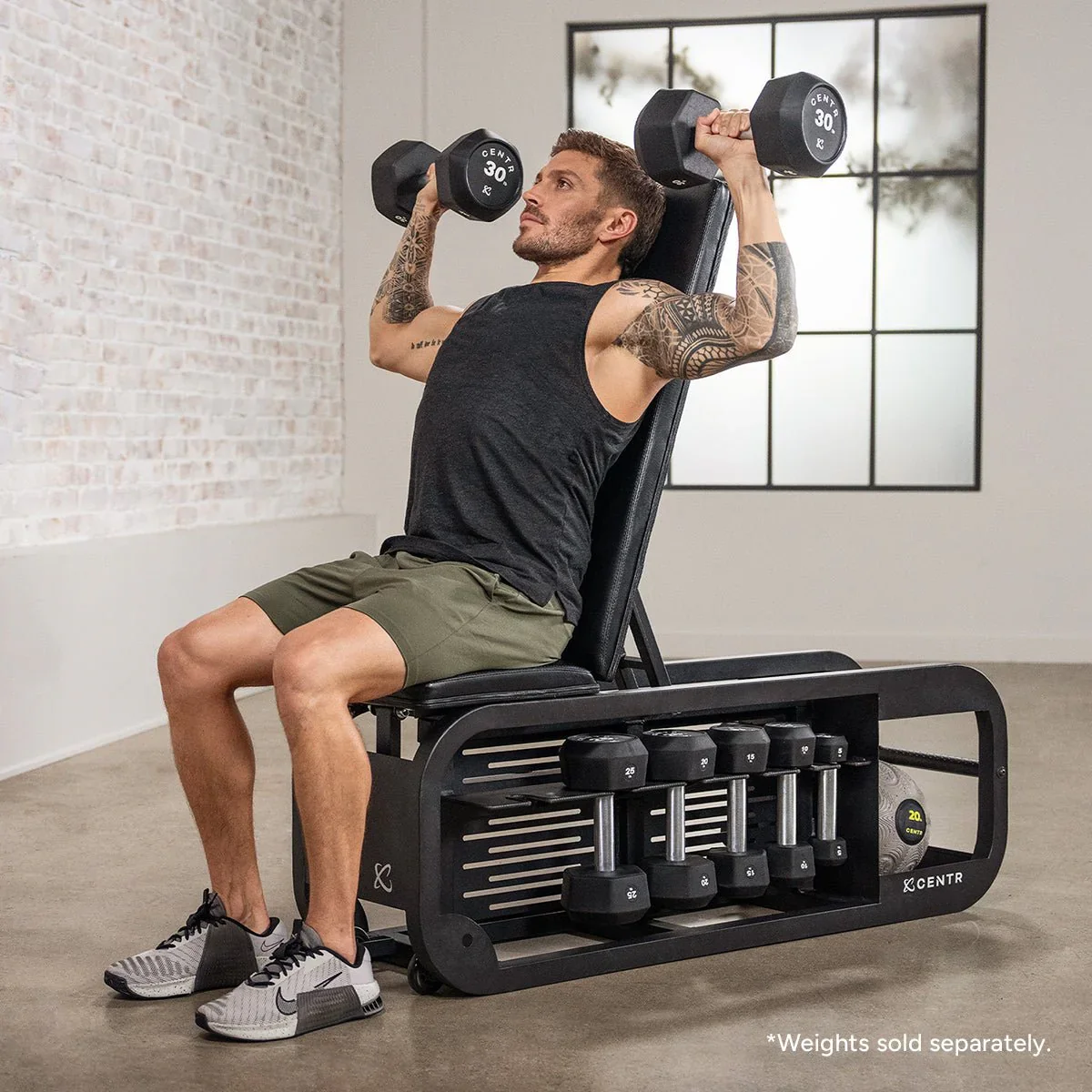 A man lifting dumbbells while seated on a multipurpose weight bench in a gym. He is wearing a black sleeveless shirt, gray shorts, and sneakers, with tattoos on his arms. The gym has a large window and a white brick wall in the background.