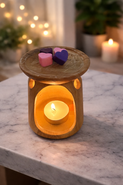 Wooden wax melter on a marble table with a lit tealight candle inside and heart-shaped wax melts on top, with soft fairy lights blurred in the background