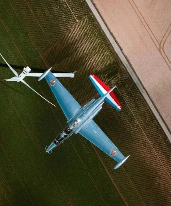 A fighter jet flying over farmland with a small wind turbine nearby.