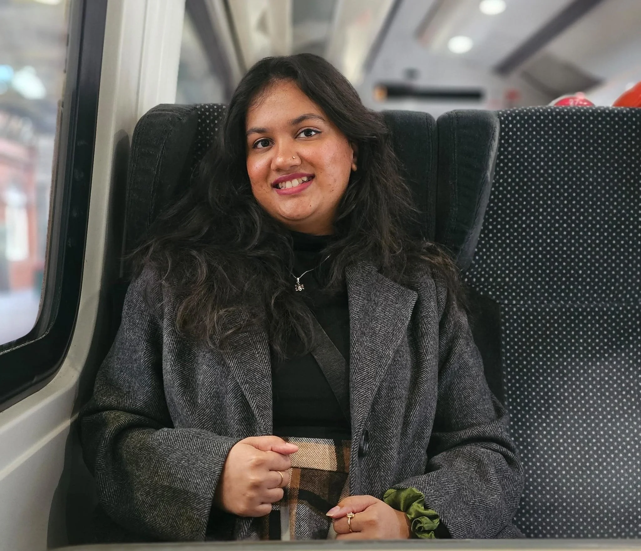 A woman with long dark hair and a smiling face sitting on a train seat with a plaid bag in her lap, wearing a gray blazer and black shirt.