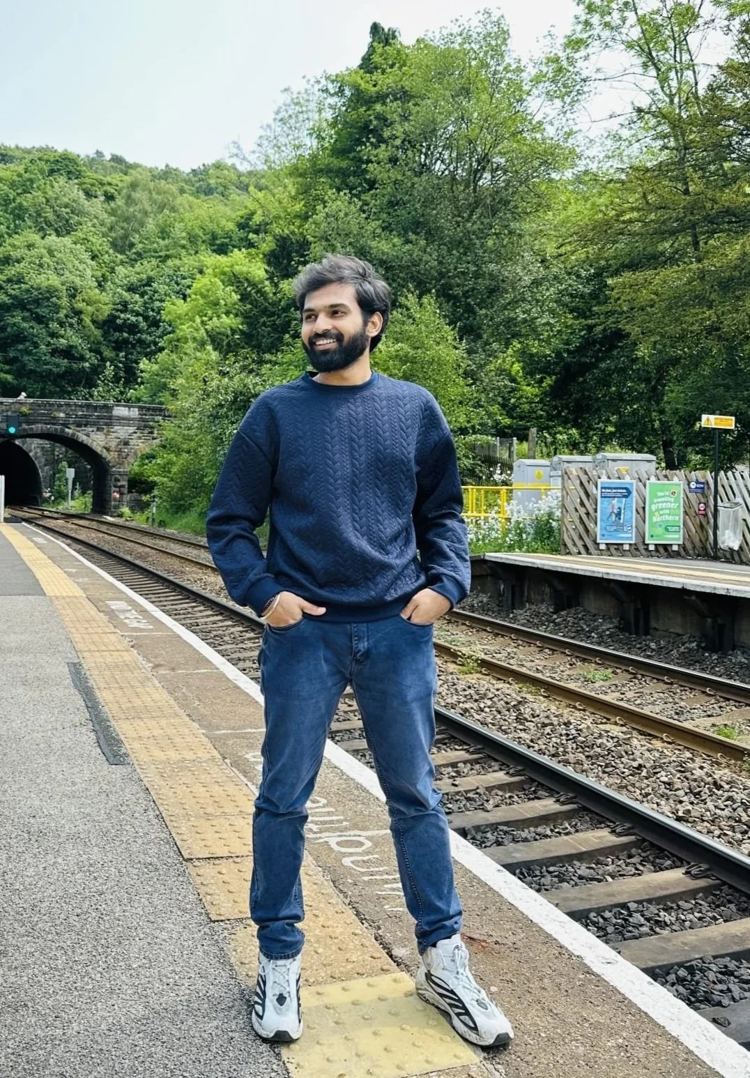 A man with a beard and dark hair, wearing a navy blue sweater, blue jeans, and white sneakers, stands on a train platform with green trees and train tracks in the background.