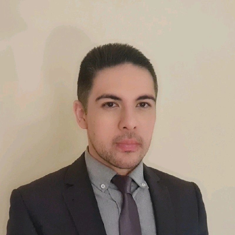 A young man in a dark suit, gray shirt, and dark tie, standing against a plain, light-colored wall.