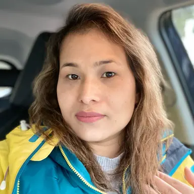 A woman with light brown hair sitting in a car, wearing a blue jacket with yellow accents.