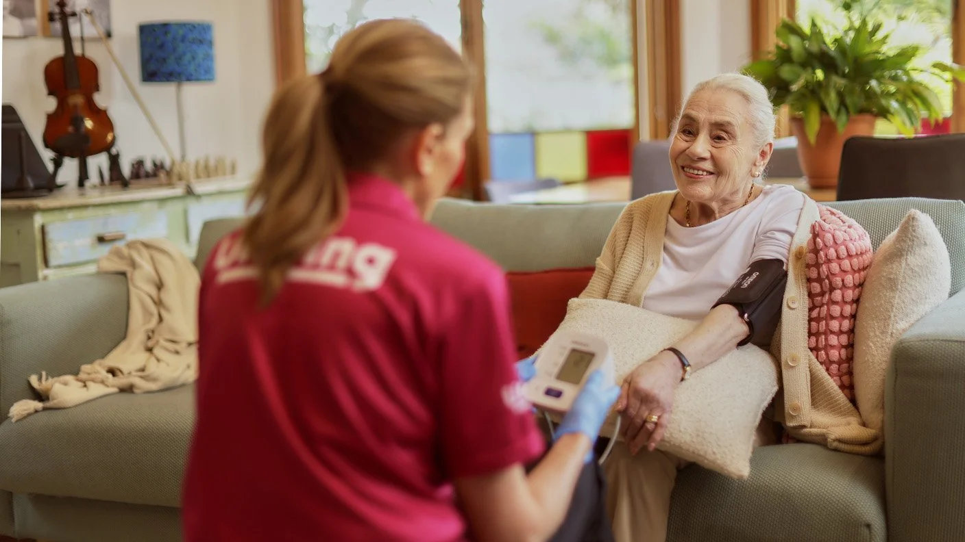 A healthcare professional taking an elderly woman's blood pressure at home. The woman is sitting on a sofa, smiling, with a pillow on her lap. The professional is wearing gloves and holding a digital blood pressure monitor. A violin and other decorative items are in the background.