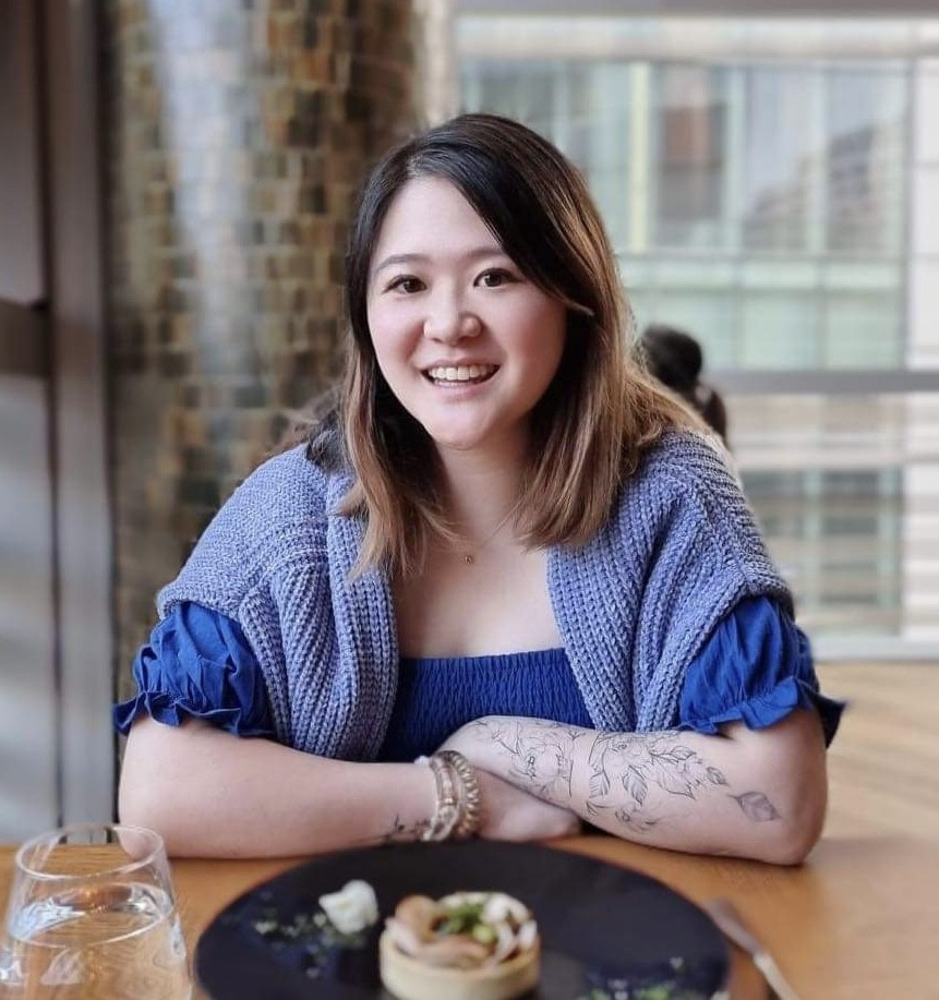 A woman with shoulder-length brown hair, smiling, wearing a blue top and a knitted vest, sitting at a table in a restaurant or cafe, with a glass of water and a small dish of food on the table.