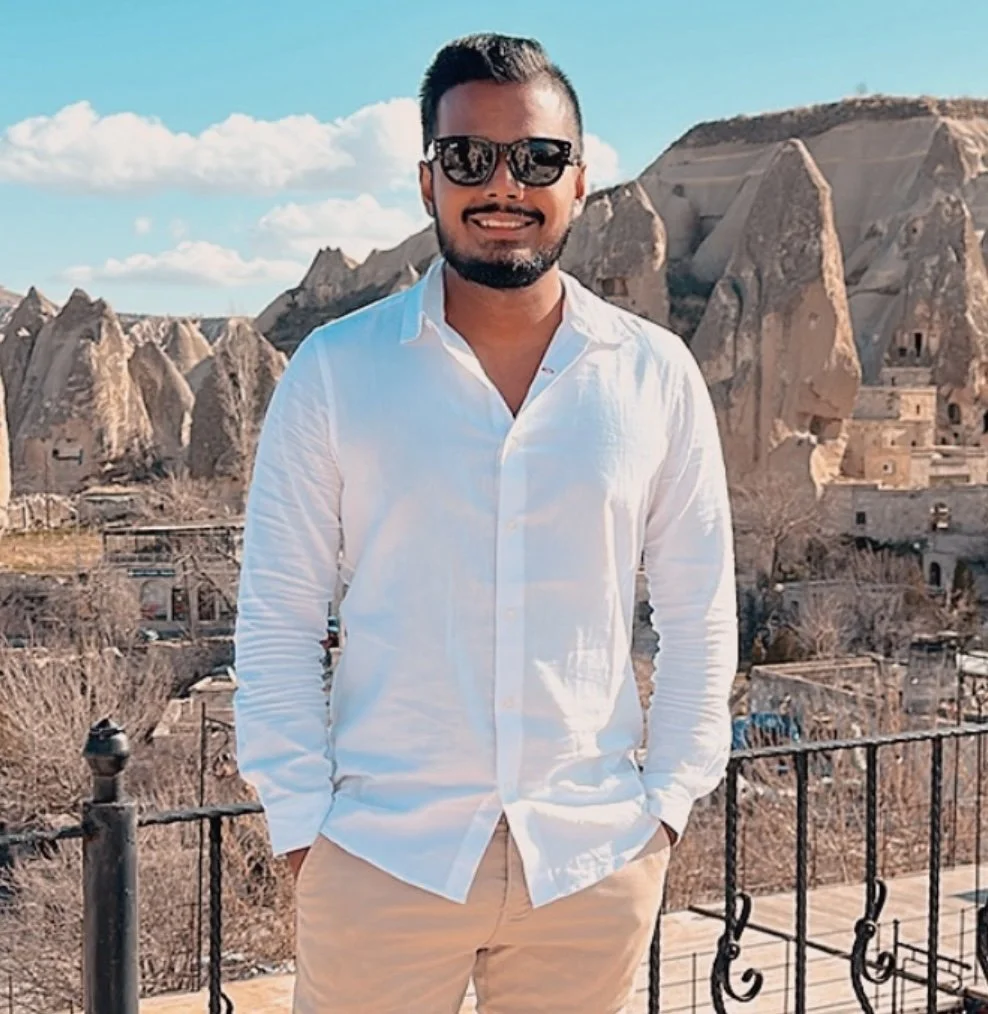 A man with dark hair, sunglasses, a white dress shirt, and beige pants standing outdoors with rock formations and cave dwellings in the background.