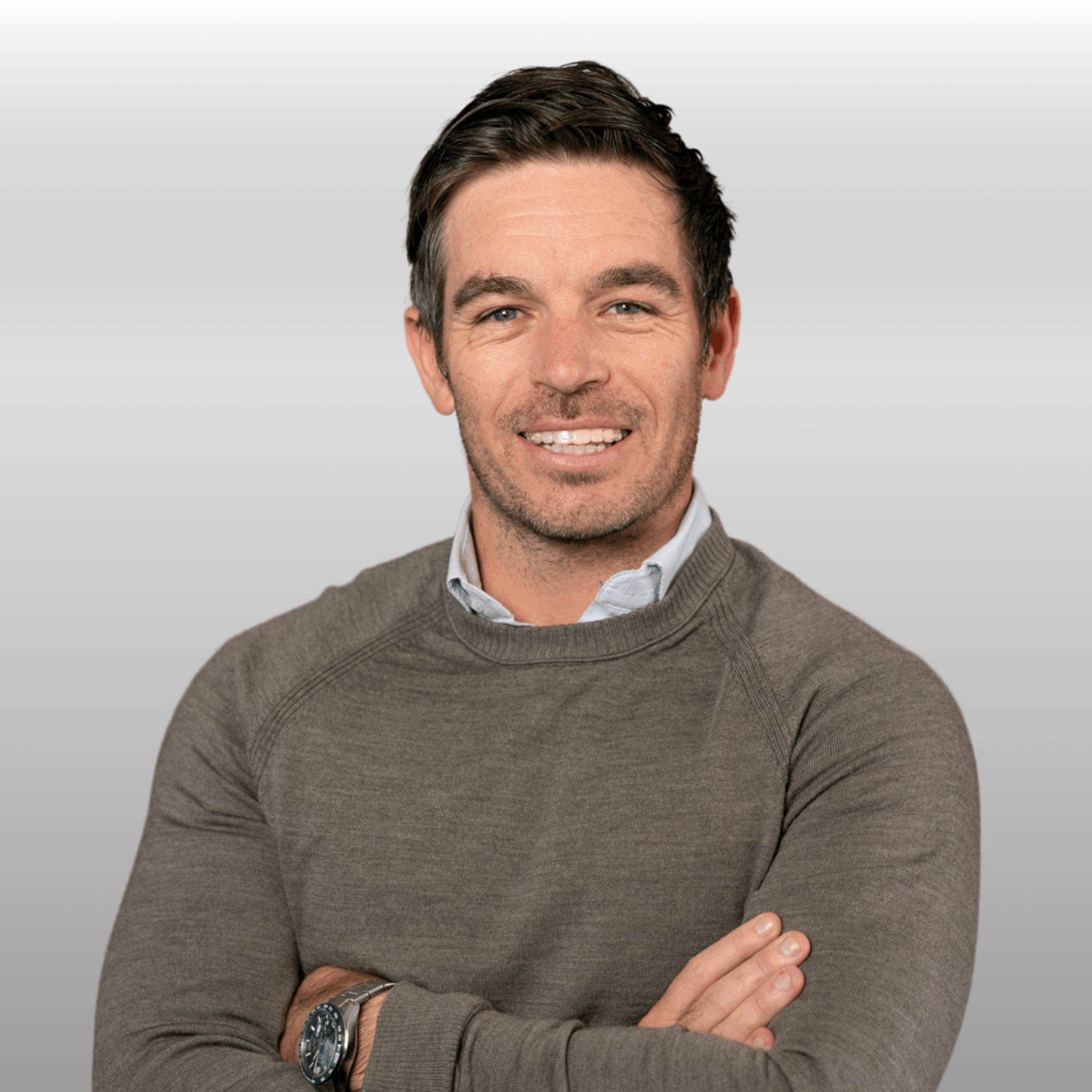 A smiling man with crossed arms, wearing a gray sweater and a watch, against a neutral background.