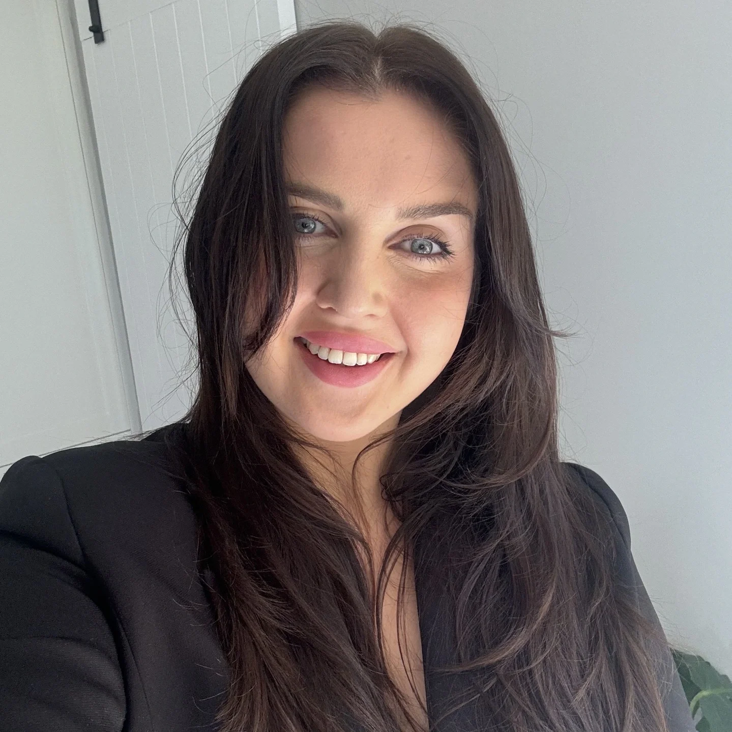 A woman with long dark hair and blue eyes smiling at the camera, dressed in a black jacket, standing indoors with a light-colored wall behind her.