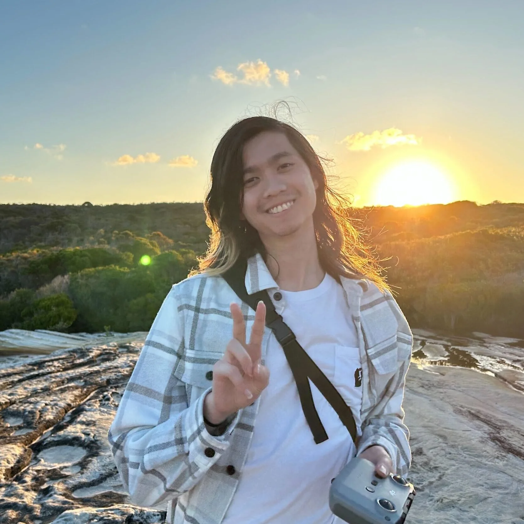 A young person with long black hair smiling and making a peace sign pose during sunset outdoors, holding a camera, with a trail and trees in the background.