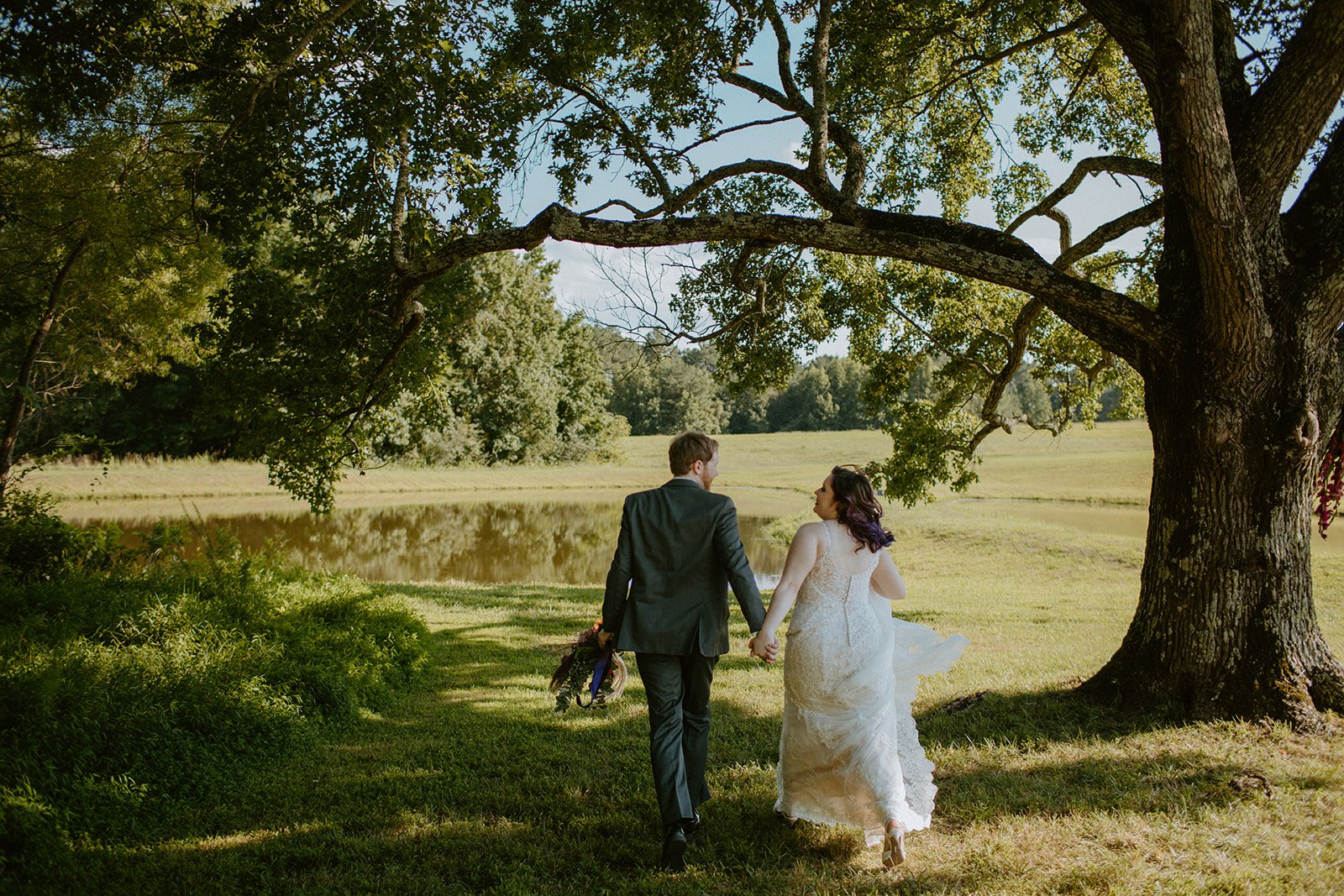 A newlywed couple, a man in a black suit and a woman in a white wedding dress, walking hand-in-hand under a large tree near a pond on a sunny day.