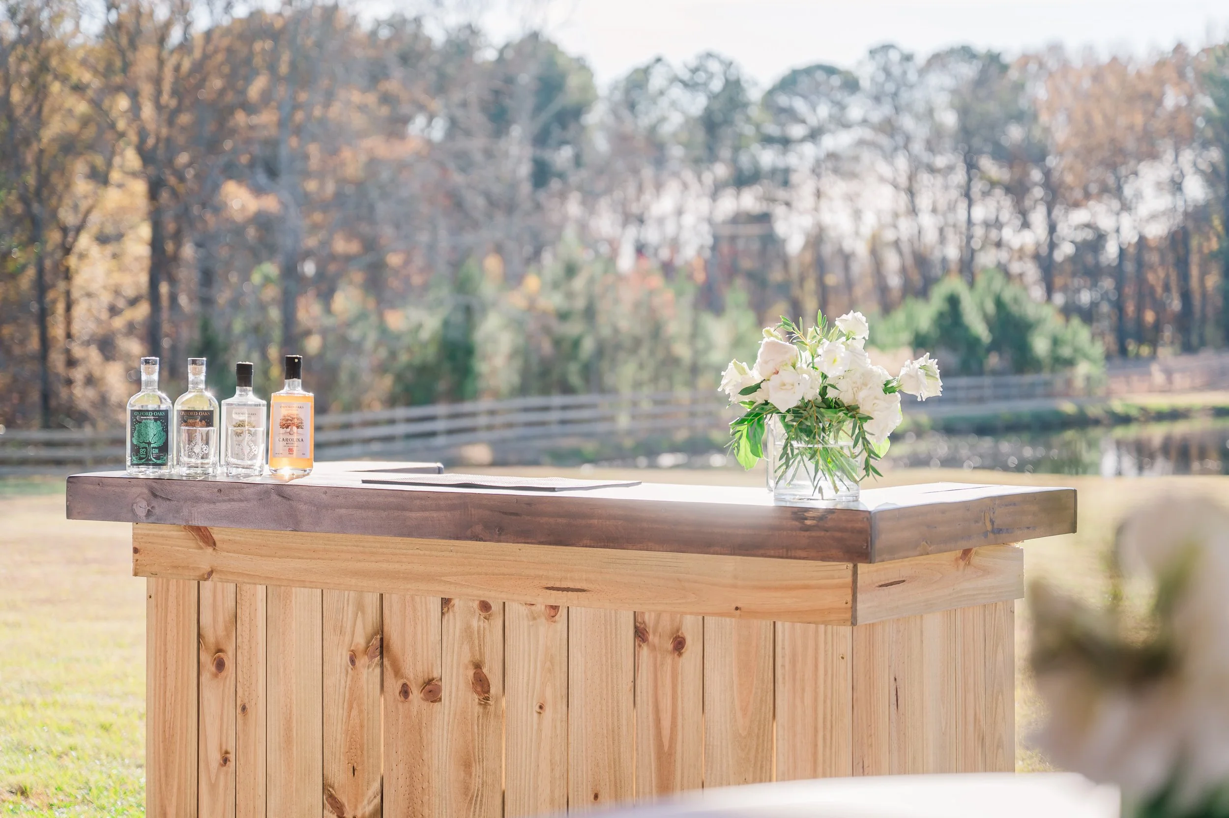 Wooden bar table outdoors with three bottles of liquor and a glass vase with white flowers. Trees and a fence are visible in the background.