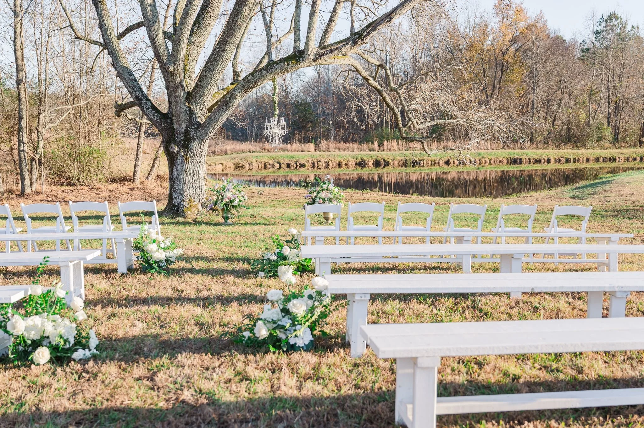 Outdoor wedding ceremony setup with white chairs and floral arrangements near a pond, large leafless trees, and a chandelier hanging from a tree branch.