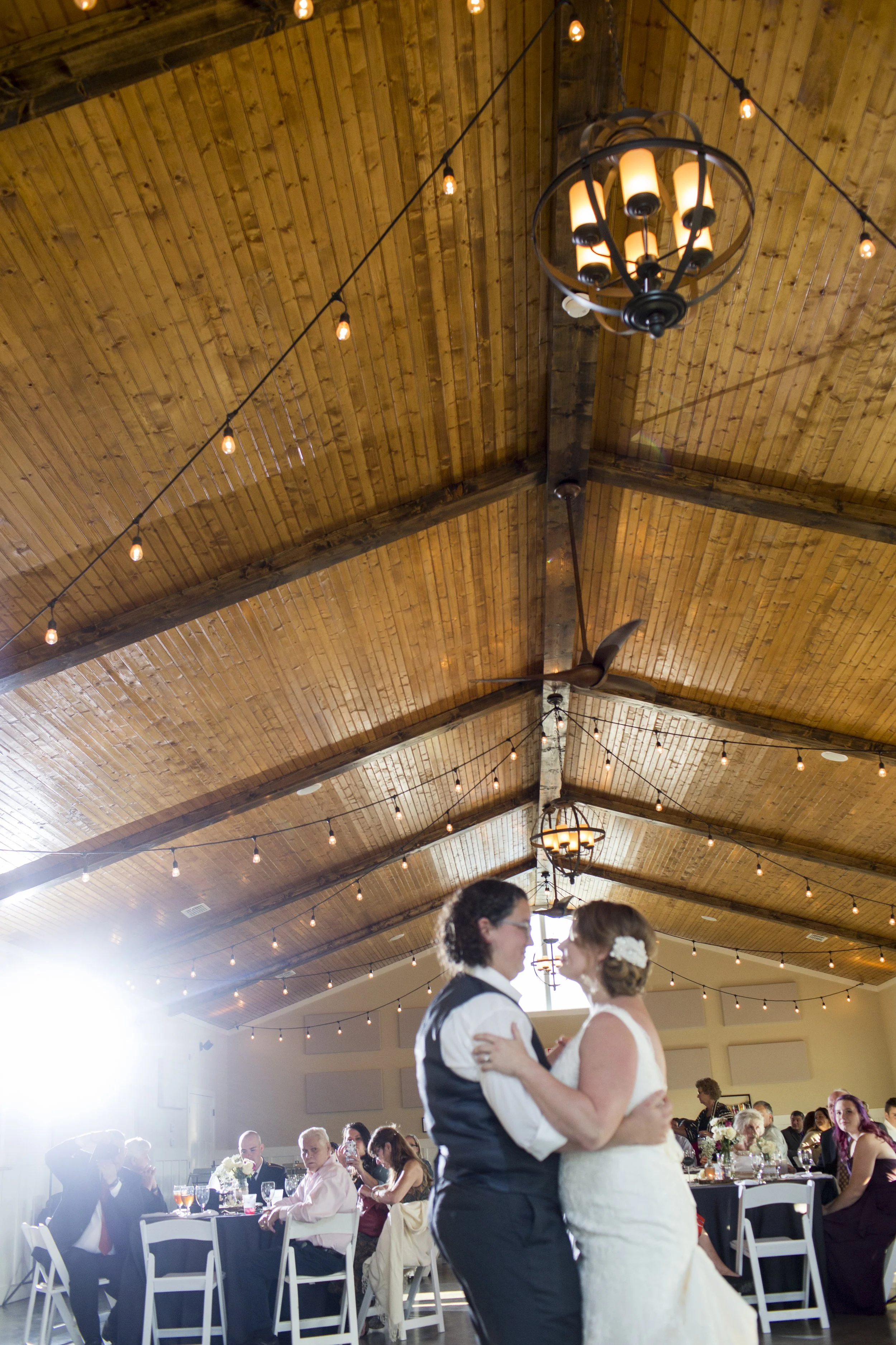 A bride and groom are dancing at their wedding reception in a decorated venue with string lights and a wooden ceiling. Guests are seated at tables watching them.