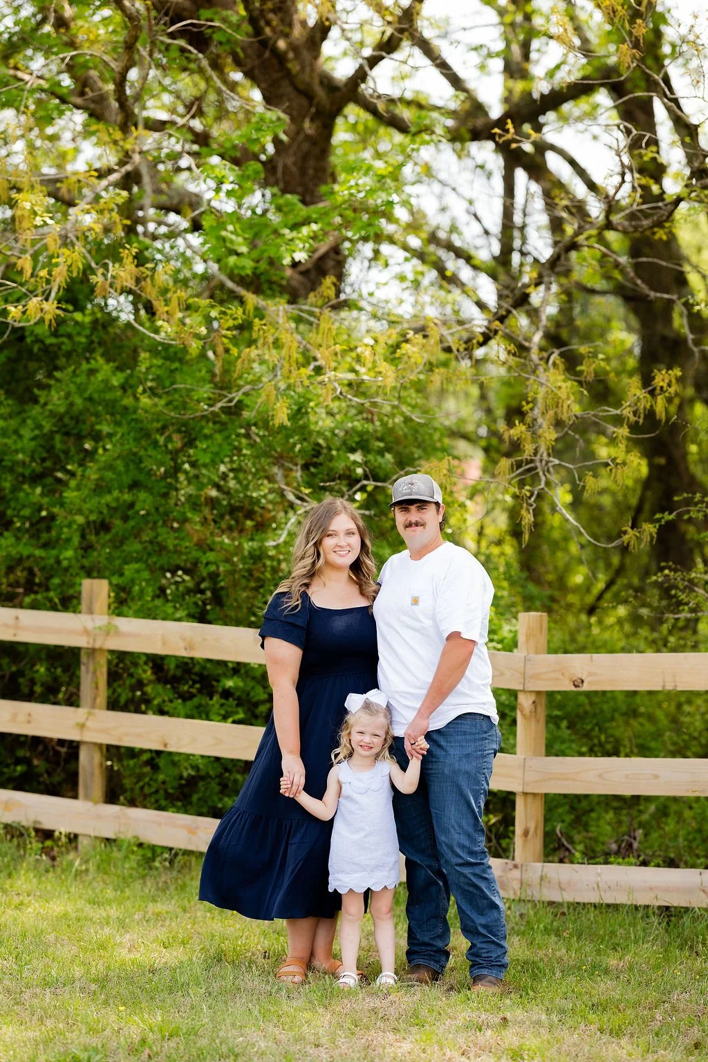 A family of three standing outdoors in front of a wooden fence and large trees, smiling and holding hands. The mother is wearing a navy blue dress, the father in a white t-shirt and cap, and the young girl in a light blue dress with a white bow in he