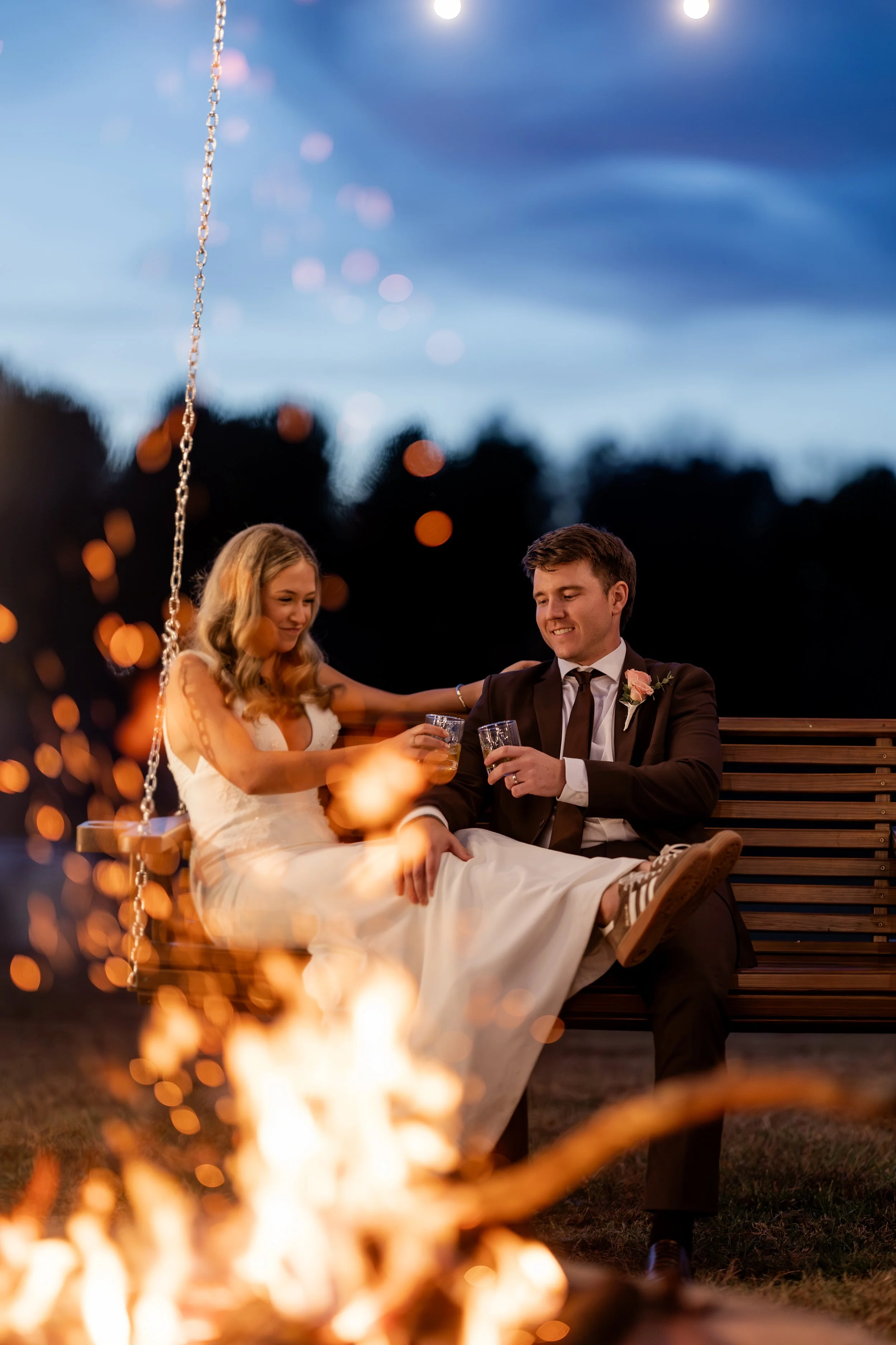 A newlywed couple sitting on a wooden bench outdoors at dusk, raising glasses for a toast, with a campfire in the foreground and string lights in the background.