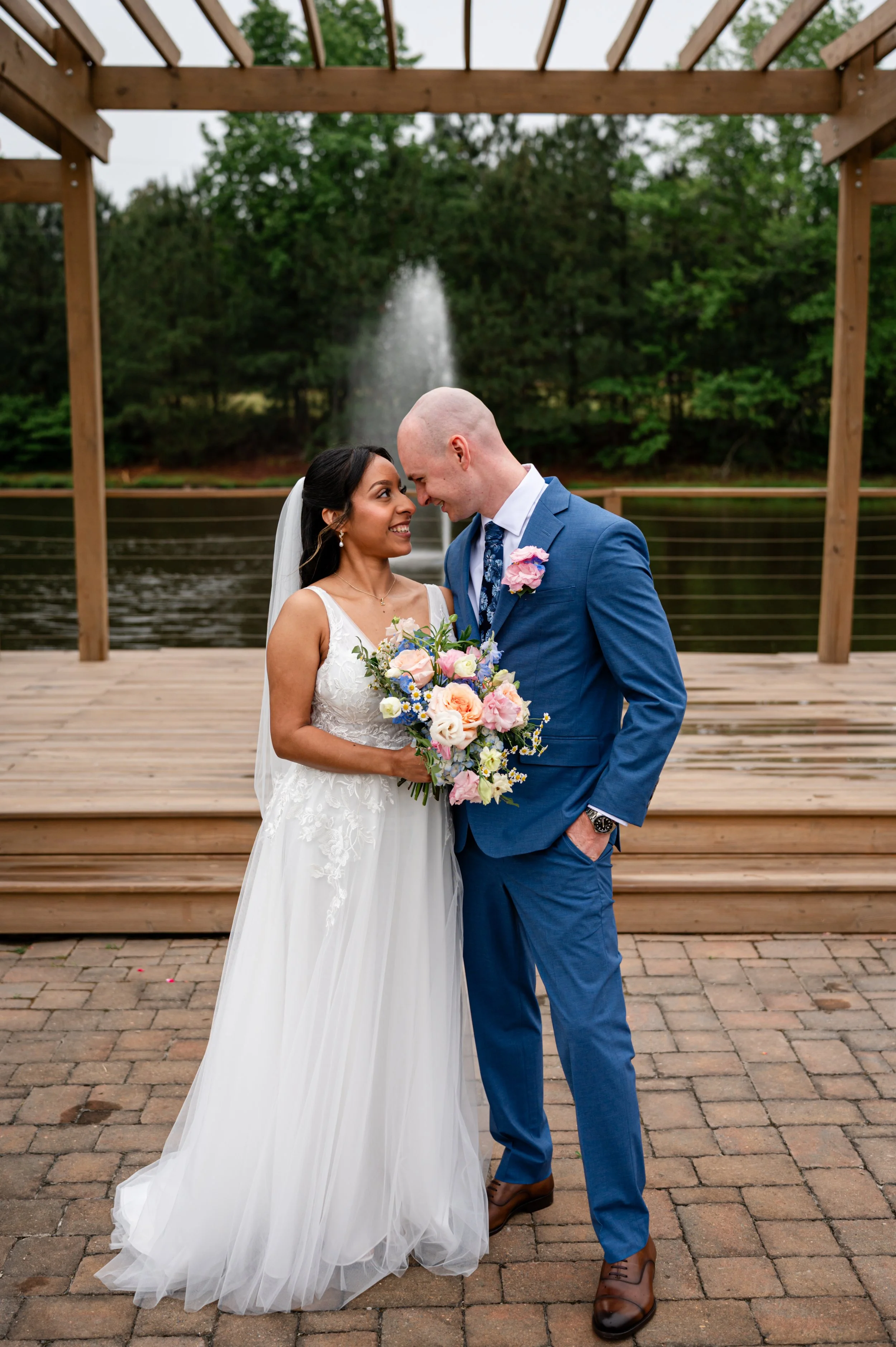 A newlywed couple is standing on a wooden deck outdoors, sharing an intimate moment. The bride is wearing a white lace wedding dress and holding a bouquet of pink, white, blue, and purple flowers. The groom is dressed in a blue suit with a white shir