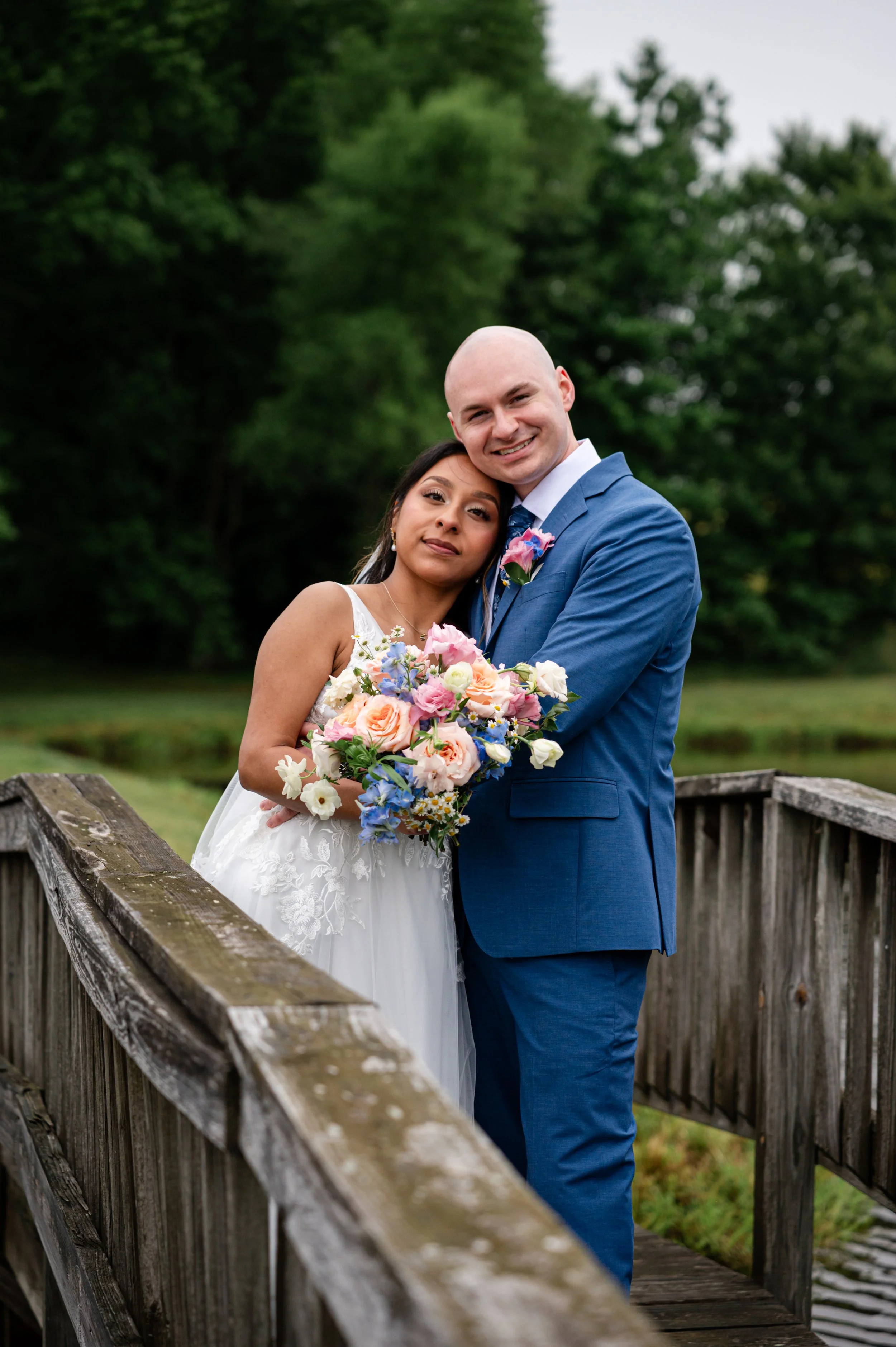 A newlywed couple standing on a wooden bridge outdoors, with lush green trees in the background. The bride is in a white wedding dress holding a bouquet, and the groom is in a blue suit with a pink boutonniere.