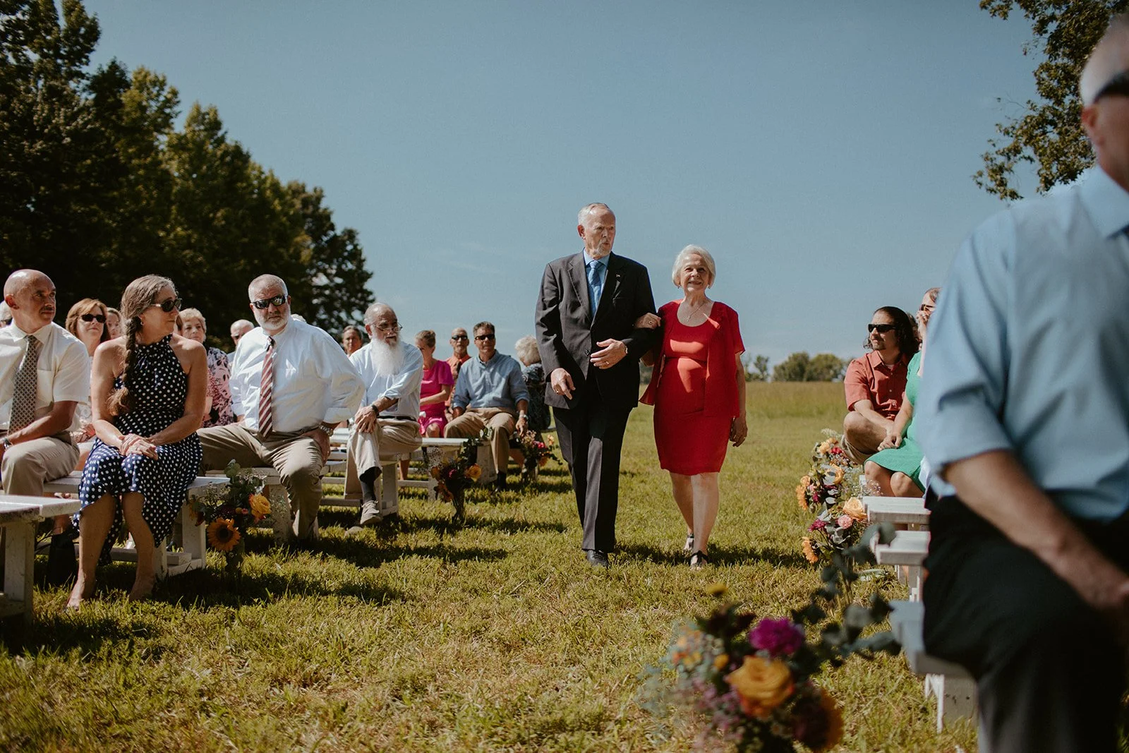 People dressed in formal and casual clothing are seated on benches outdoors on a sunny day, attending a ceremony or gathering. Some individuals are walking down an aisle between the benches.