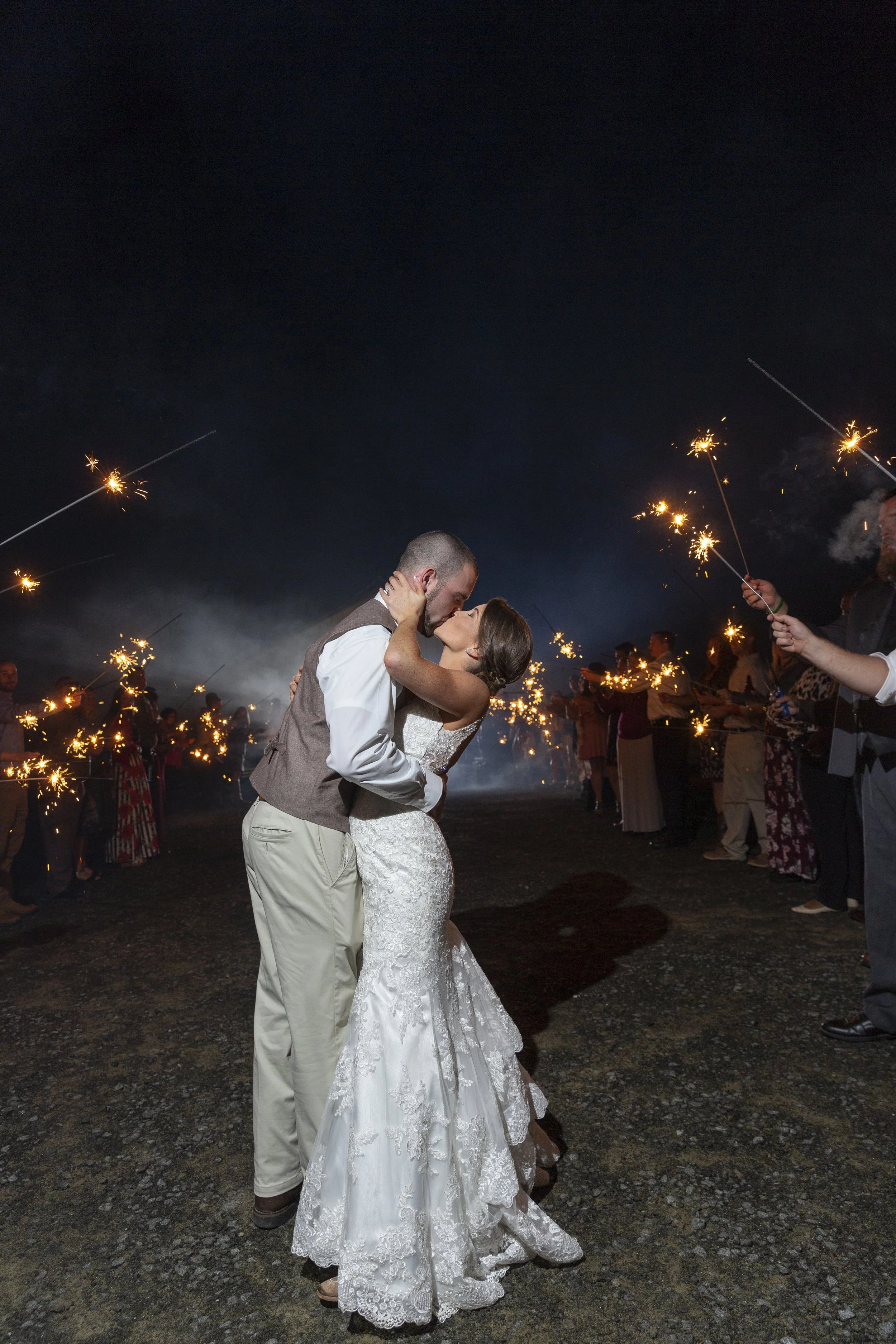 A newlywed couple sharing a kiss during their wedding celebration at night, surrounded by guests holding sparklers.