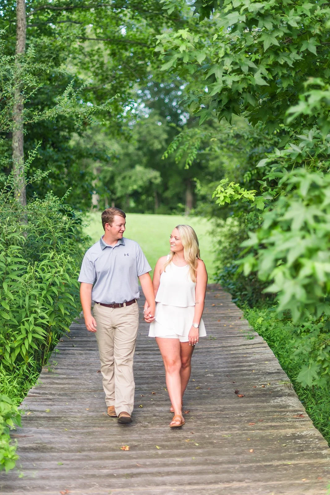 A young couple holding hands and walking on a wooden path through lush green trees and bushes, smiling at each other.