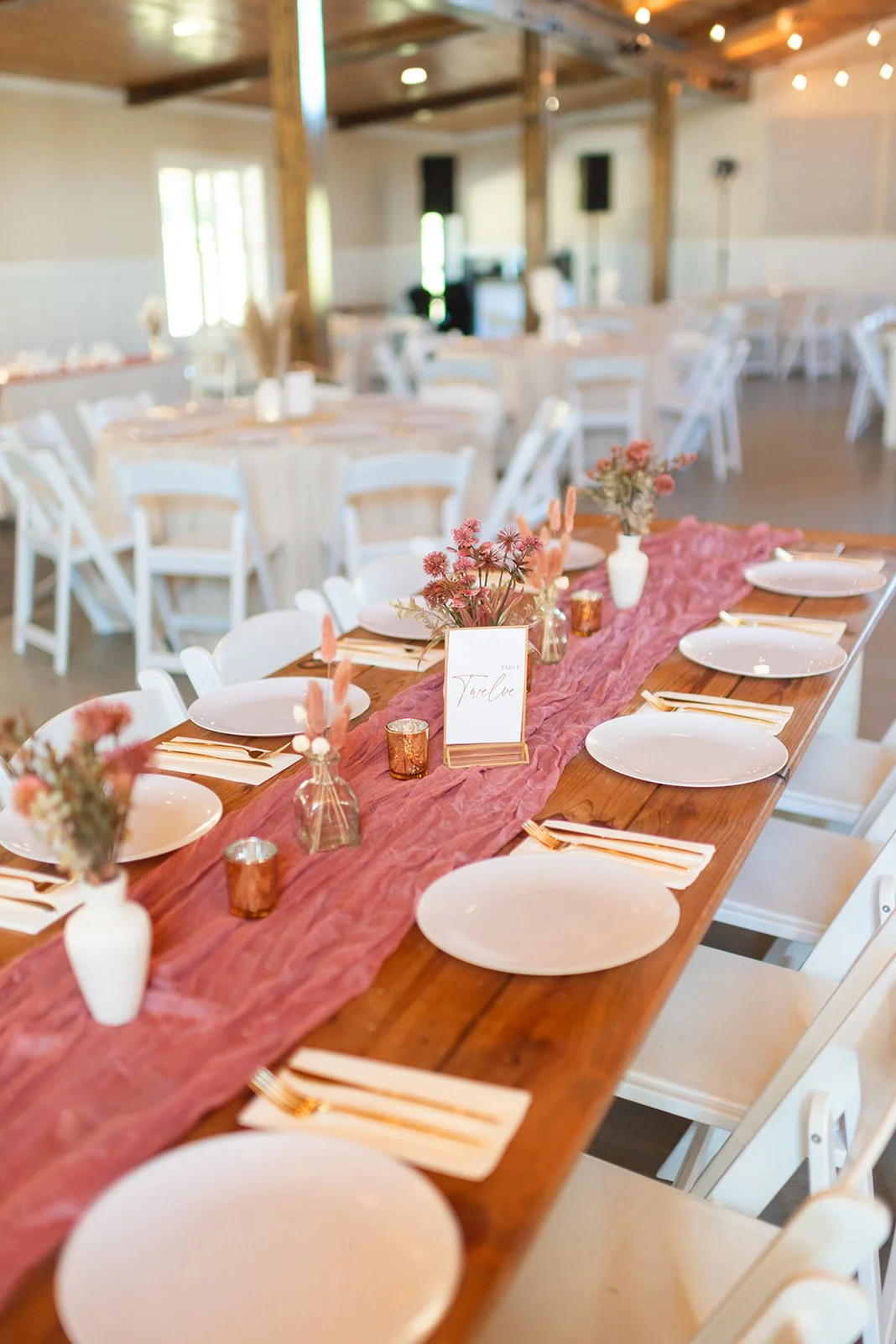 A decorated wedding reception table with a pink table runner, white plates, gold cutlery, small vases with pink flowers, and a table number sign, in a charming venue in close proximity to the north carolina triangle