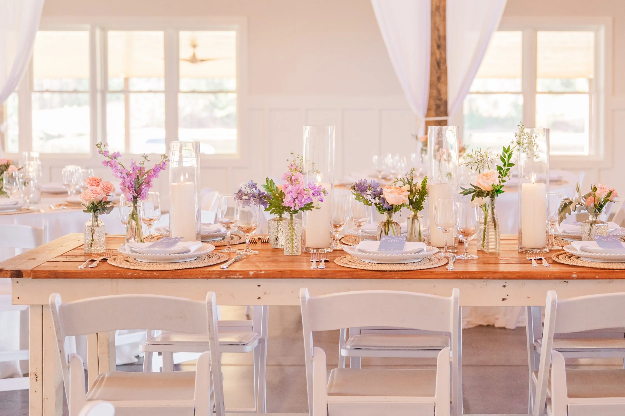 A dining table set for a celebration with floral centerpieces and candles in a bright room with large windows.