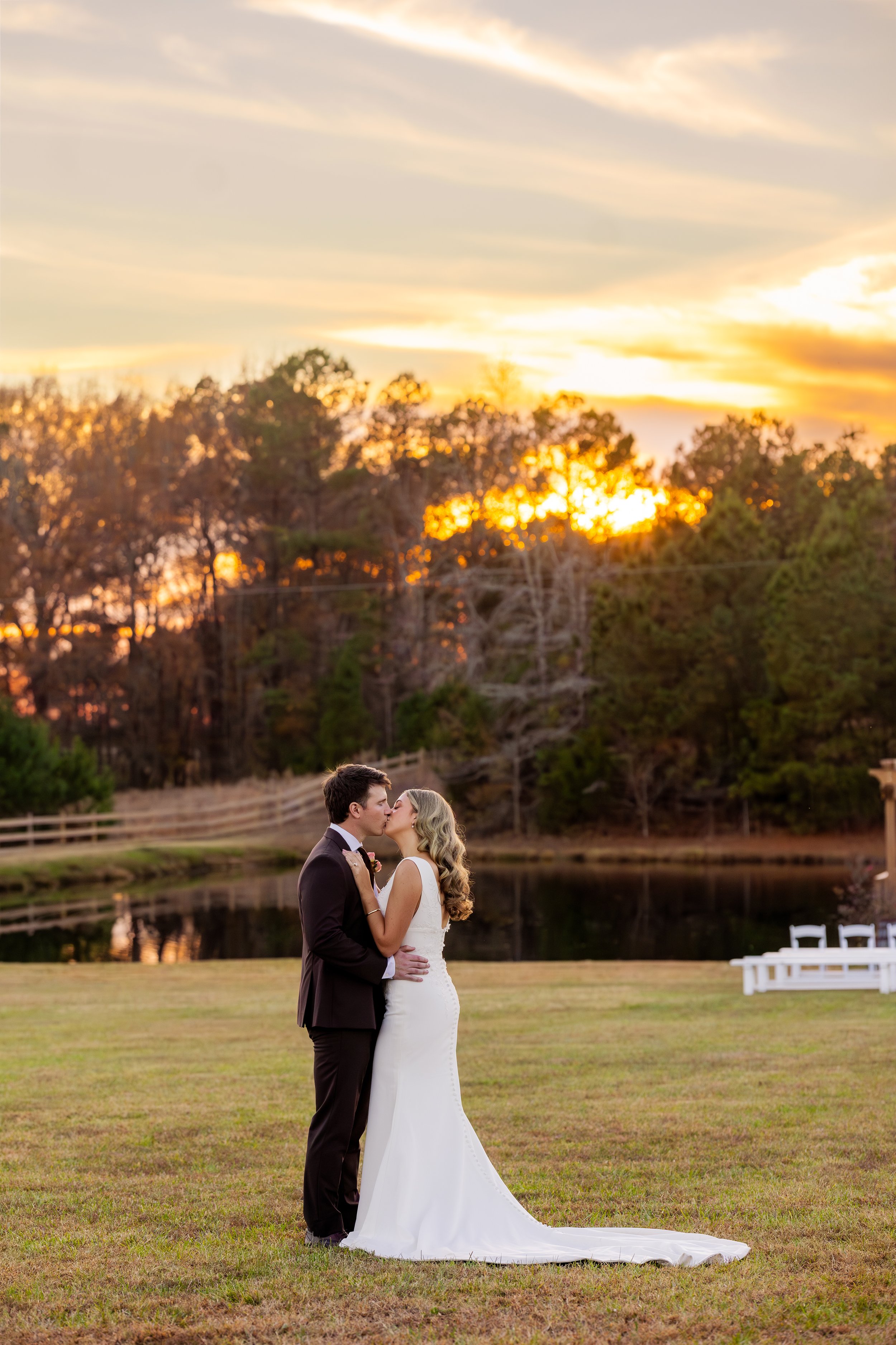 A bride and groom share a kiss during their wedding photoshoot outdoors at sunset, with trees and a pond in the background.
