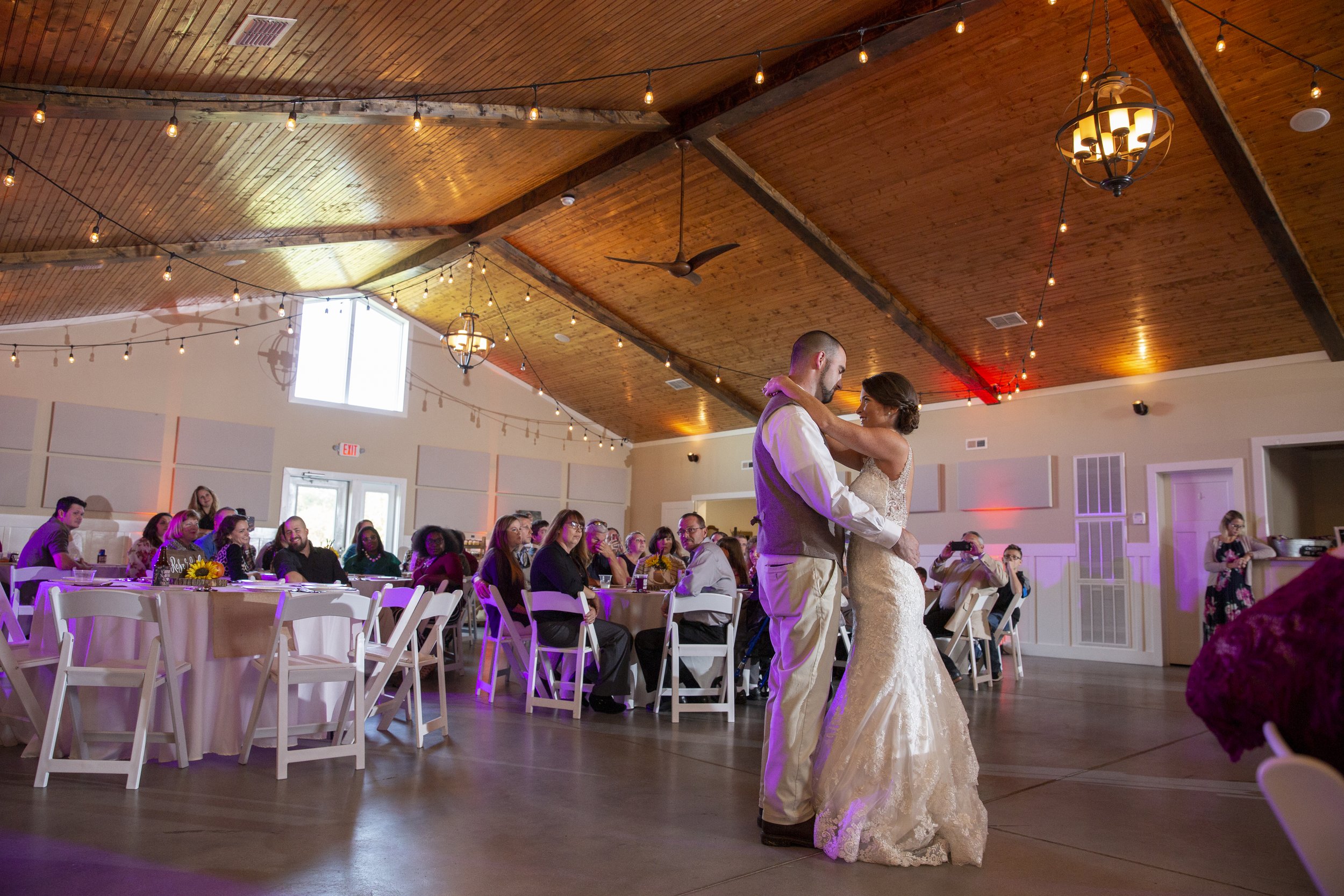 A bride and groom sharing their first dance at a wedding reception in a decorated venue with wooden ceiling, string lights, chandeliers, and seated guests.