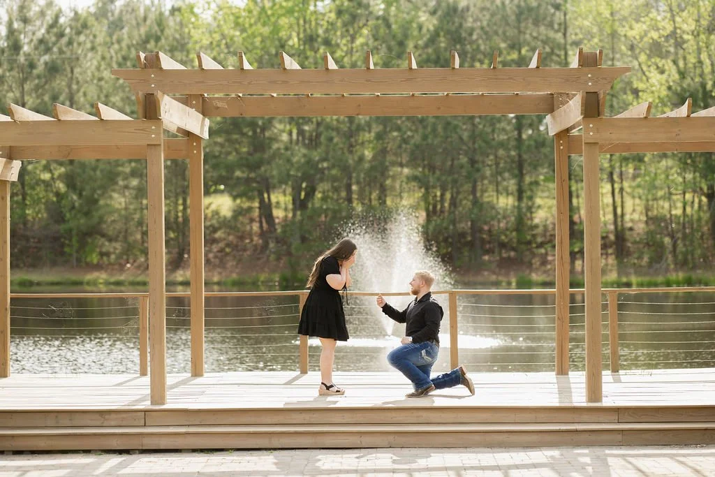 A man kneeling on one knee proposing to a woman on a wooden dock near a lake, with water splashing as he presents a ring, surrounded by trees and a wooden pergola with decorative spikes.