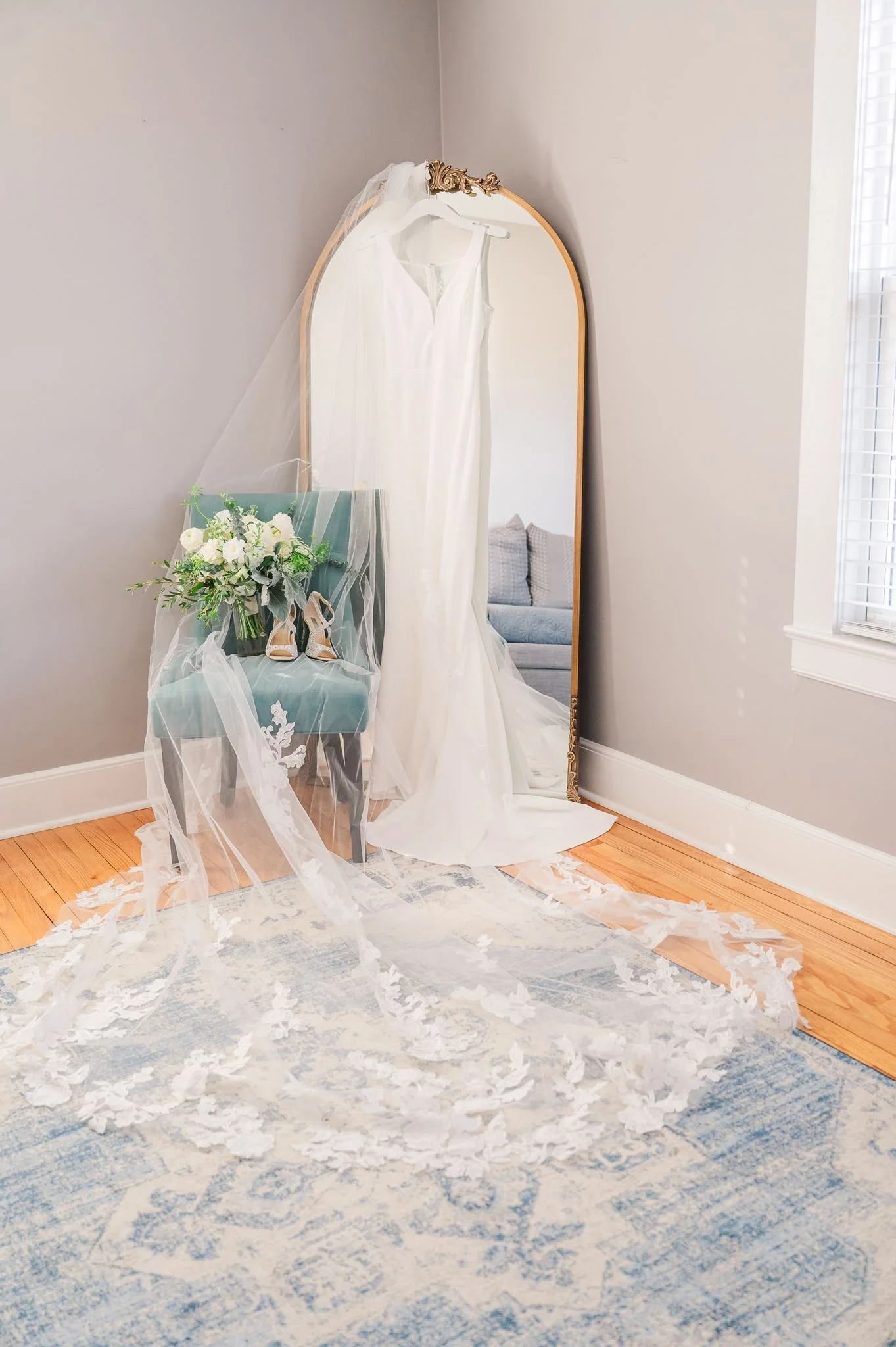 A wedding dress hanging on a mirror in a room with a bouquet of white roses and beige heels on a chair draped with tulle fabric. The room has light gray walls, a window with white shutters, and a blue and white area rug on hardwood floor.
