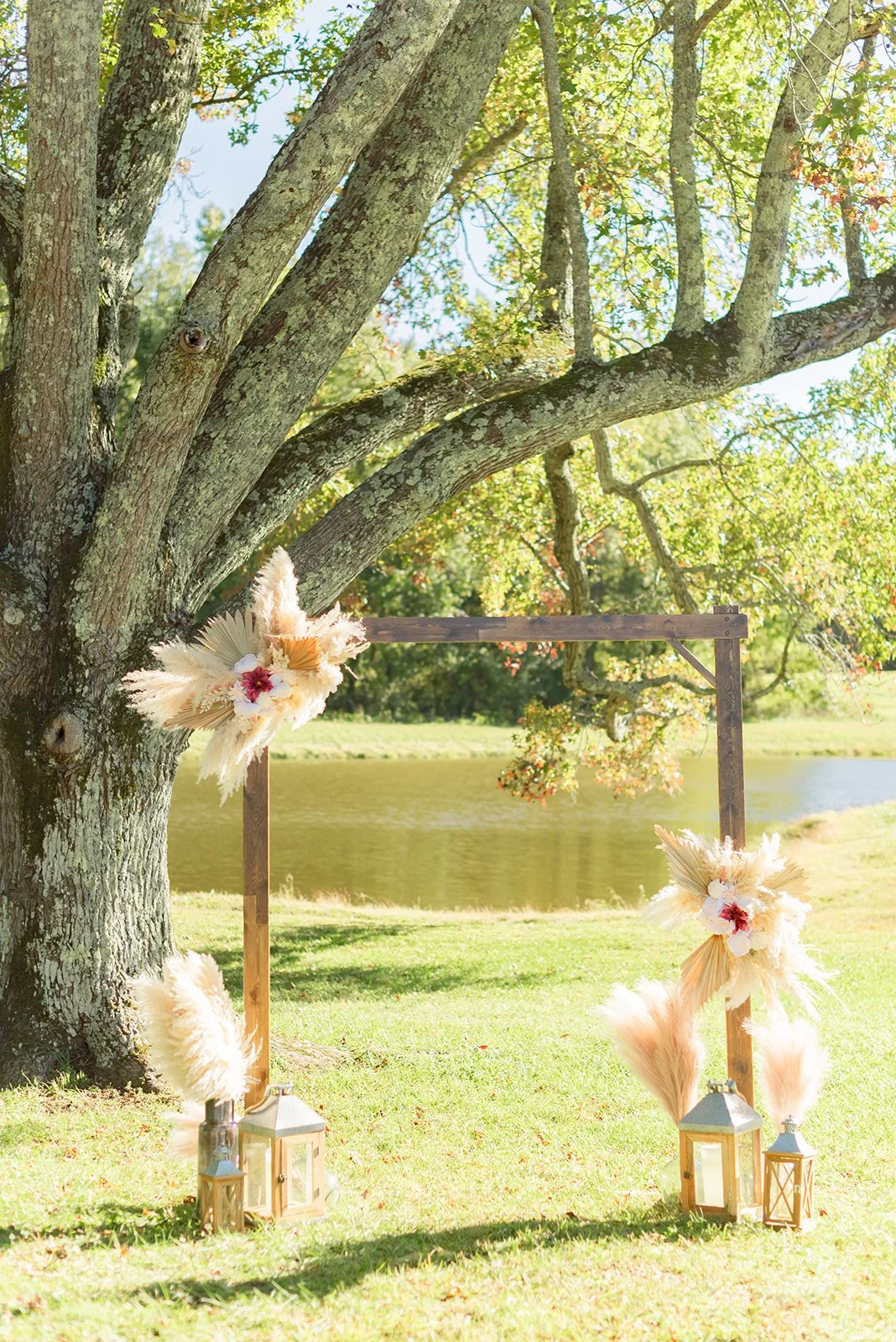 Outdoor wedding ceremony setup with a wooden arch decorated with dried flowers and pampas grass, situated next to a large tree near a pond