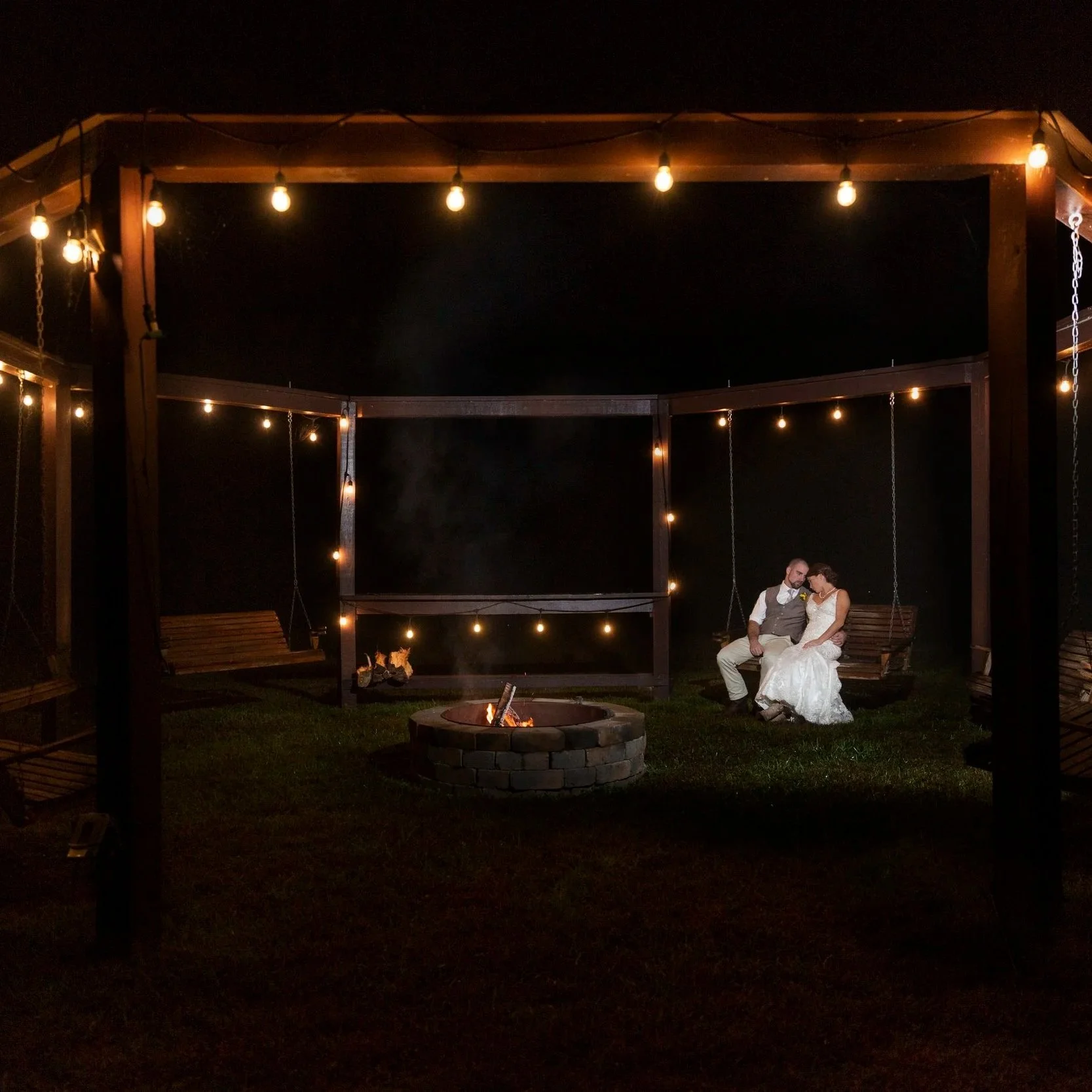 A bride and groom sitting together on a swing at night under string lights, around a fire pit in a backyard. The groom is dressed in a vest and shirt, and the bride is wearing a white wedding dress.