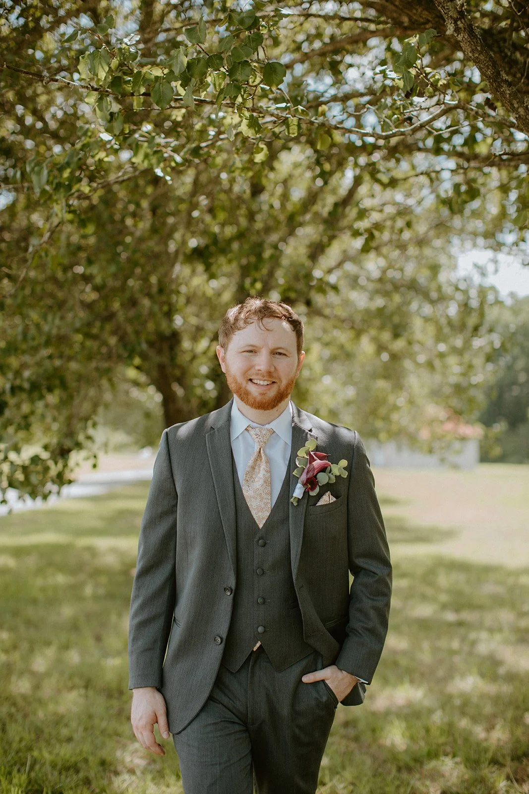 A smiling man in a gray suit with a boutonniere stands outdoors under a tree on a sunny day.