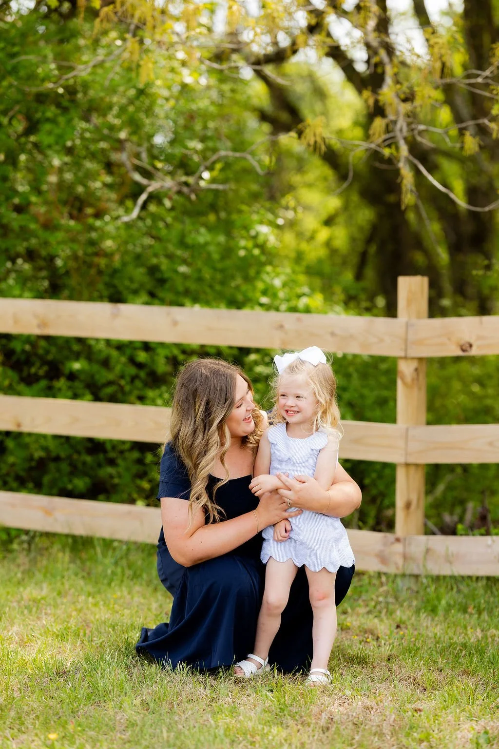 A woman with long wavy hair in a navy blue dress is kneeling on the grass, smiling at a young girl with blonde hair wearing a light blue ruffled dress and a white bow in her hair, as they share a joyful moment outdoors near a wooden fence with green 