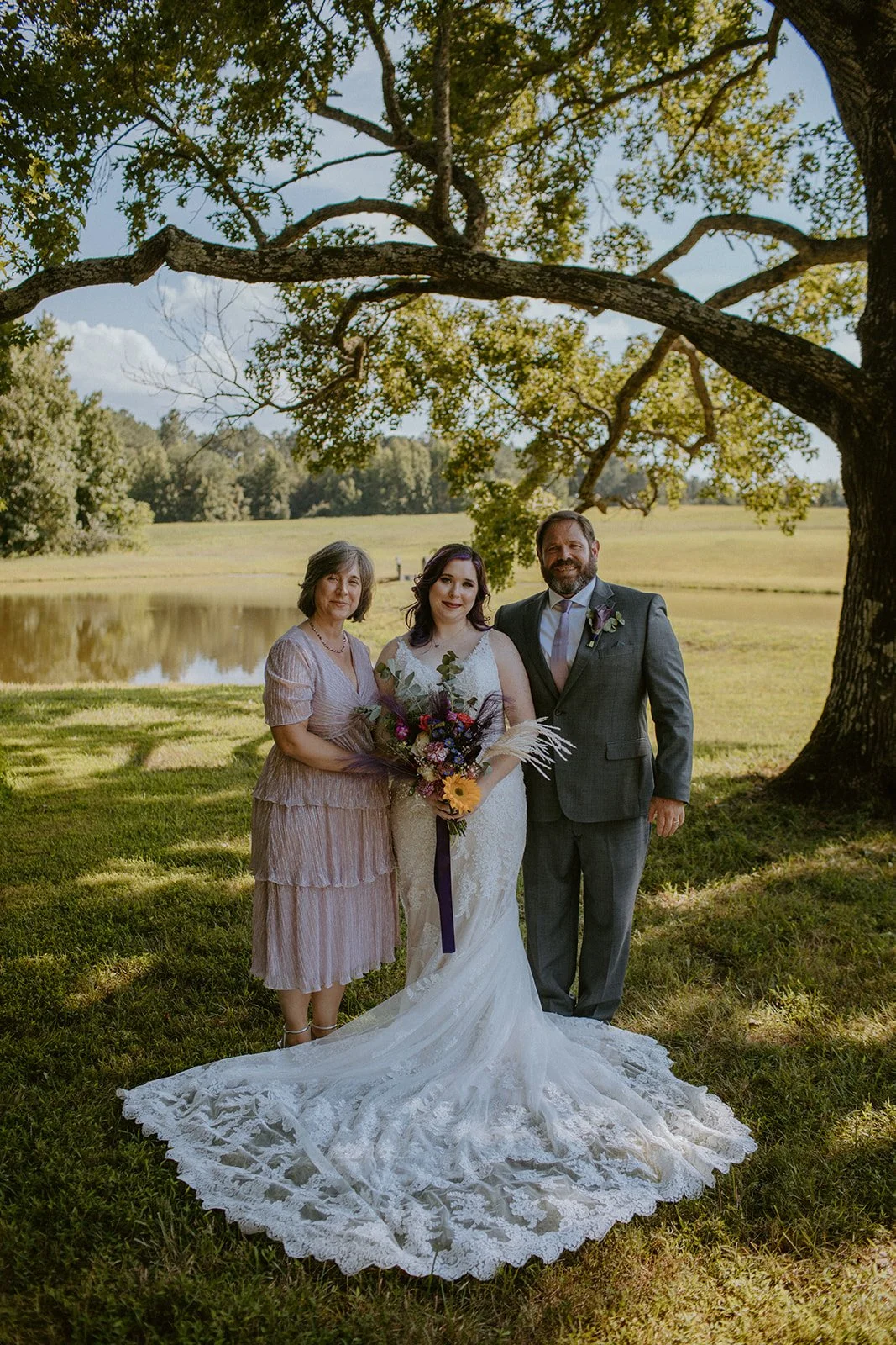 Bride in a white lace wedding gown holding a bouquet, standing between a woman in light pink dress and a man in grey suit with a tie, under a large tree near a pond, on a sunny day.