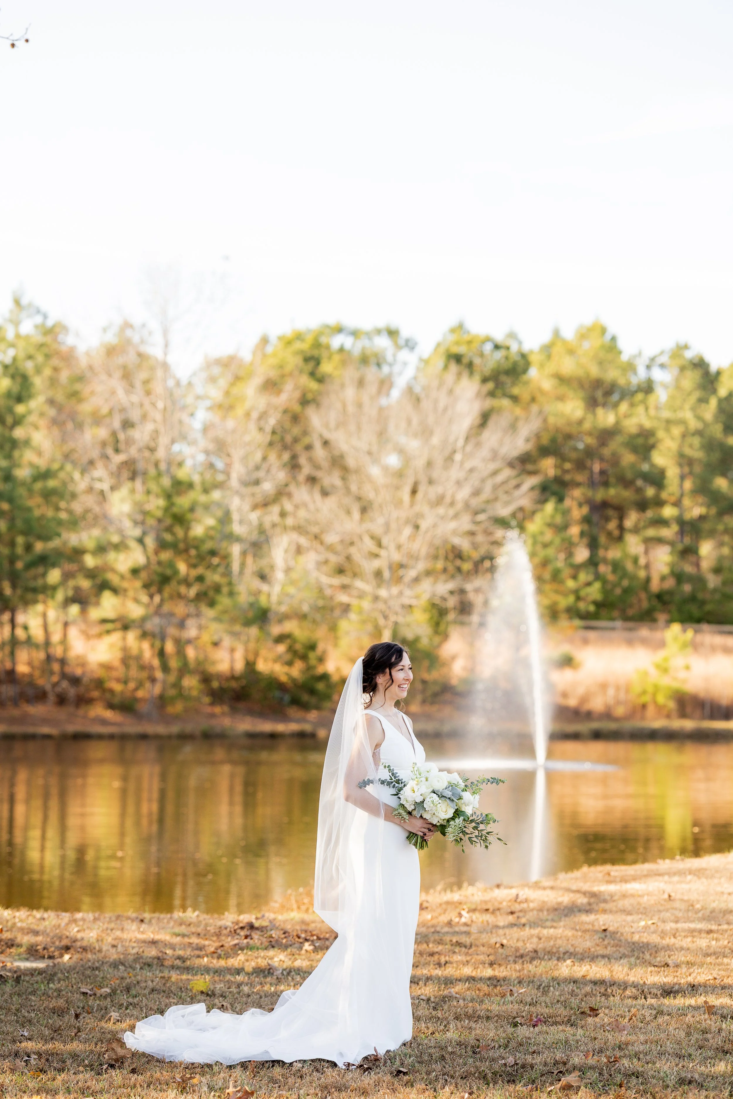 A bride in a white wedding dress holding a bouquet of white flowers standing by a pond with a fountain in the background, surrounded by autumn trees.