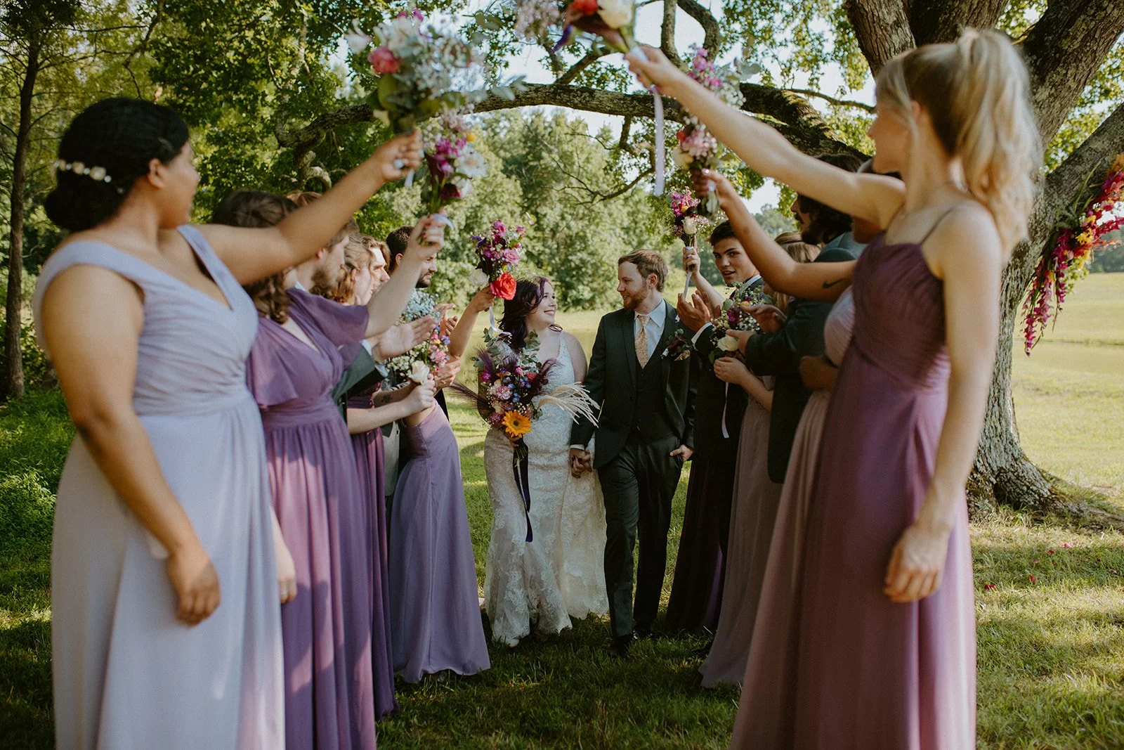 A bride and groom walking hand in hand through a floral archway, surrounded by bridesmaids and groomsmen holding bouquets, outdoors under the ceremony tree at sable and vine