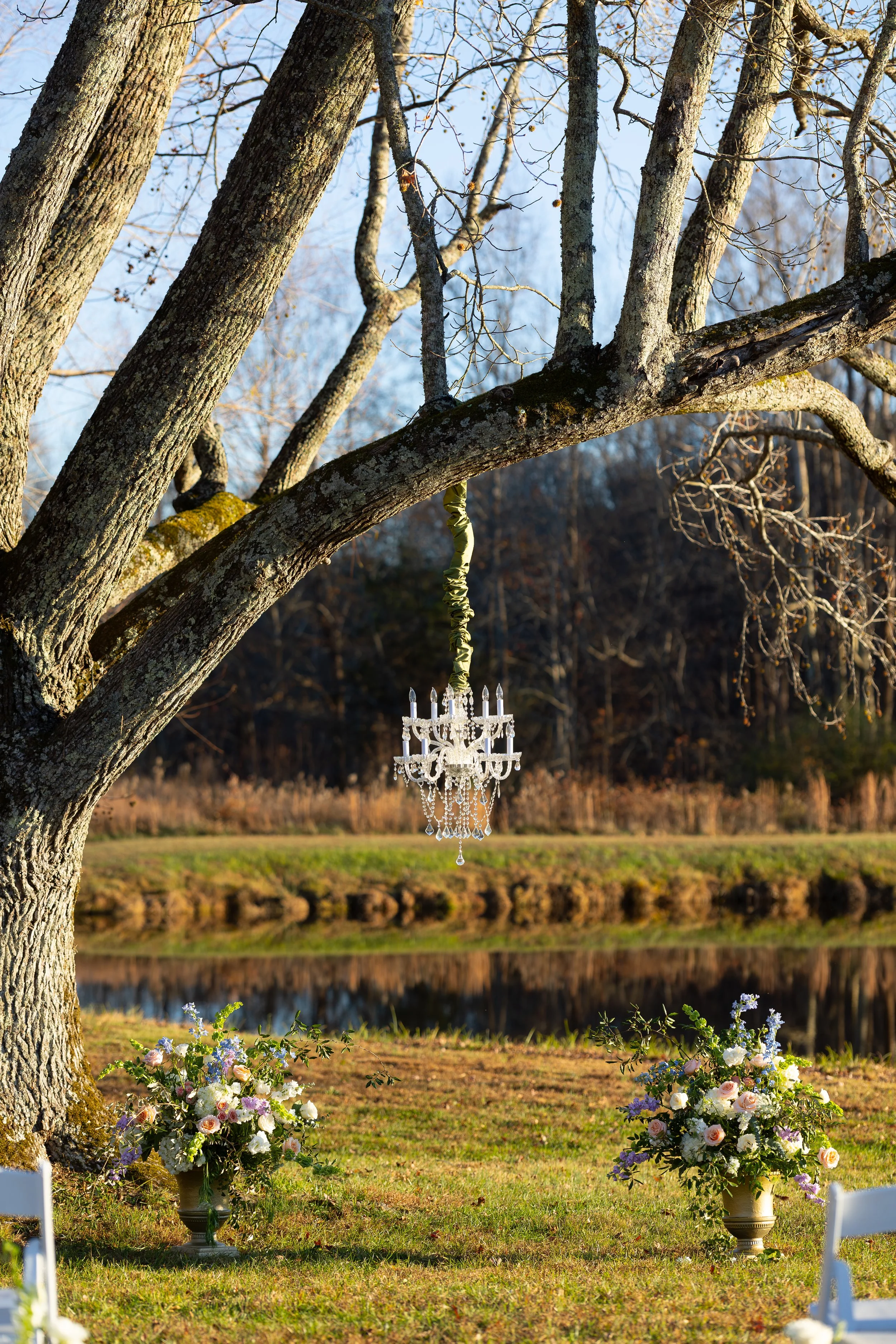 A tree with a chandelier hanging from one of its branches. There are flower arrangements in vases on the ground beneath the tree, and a fenced area nearby.