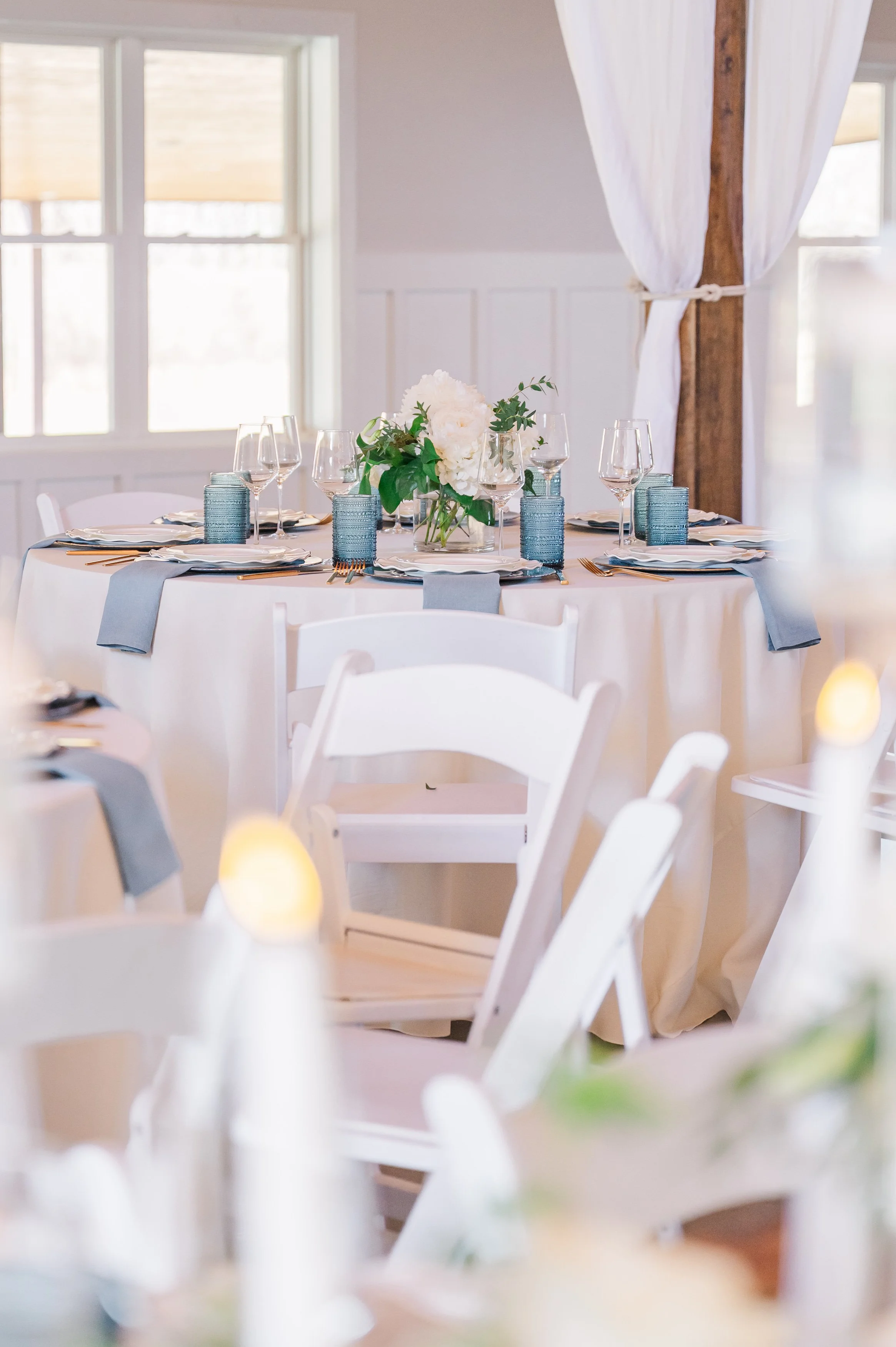 A round table decorated with a white tablecloth, a floral centerpiece with white flowers and green leaves, and place settings with gold utensils and blue glasses. The setting appears to be in a bright, rustic room with large windows, white walls, and