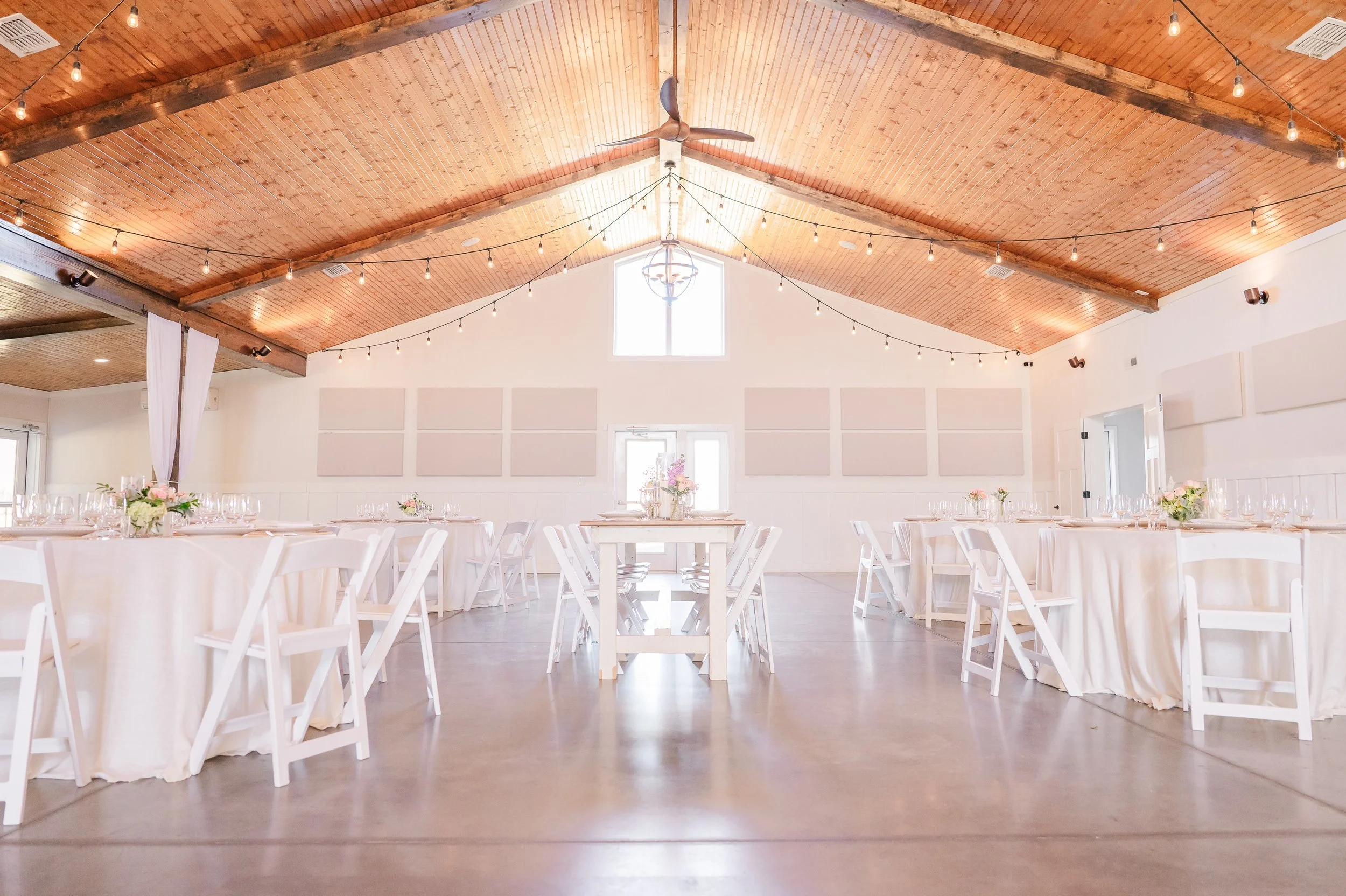 Indoor event space with high wooden ceiling, string lights, and white tables and chairs decorated with pink and white flowers.
