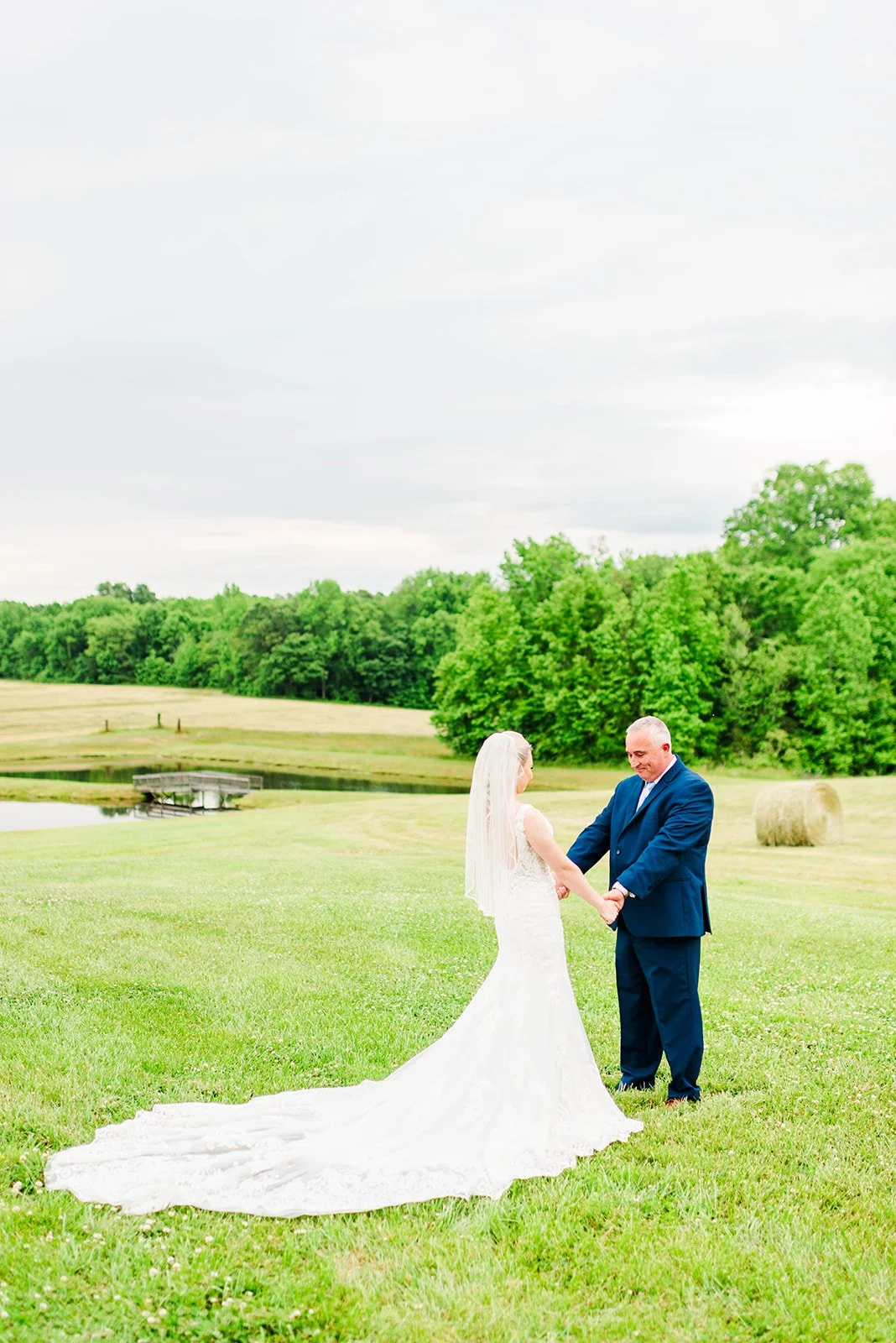 Bride and groom holding hands in a grassy field with trees and hay bales, overcast sky.