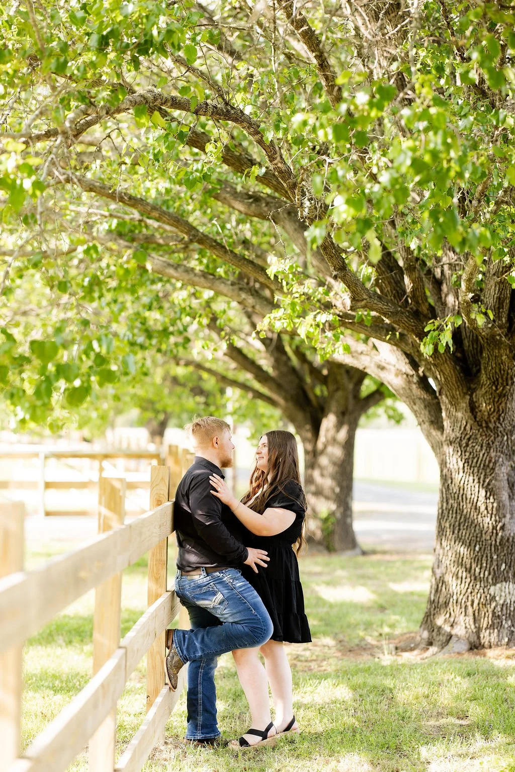 A couple with light skin and long hair standing close to each other under a large leafy tree, with the woman touching the man's shoulder and the man leaning against a wooden fence.