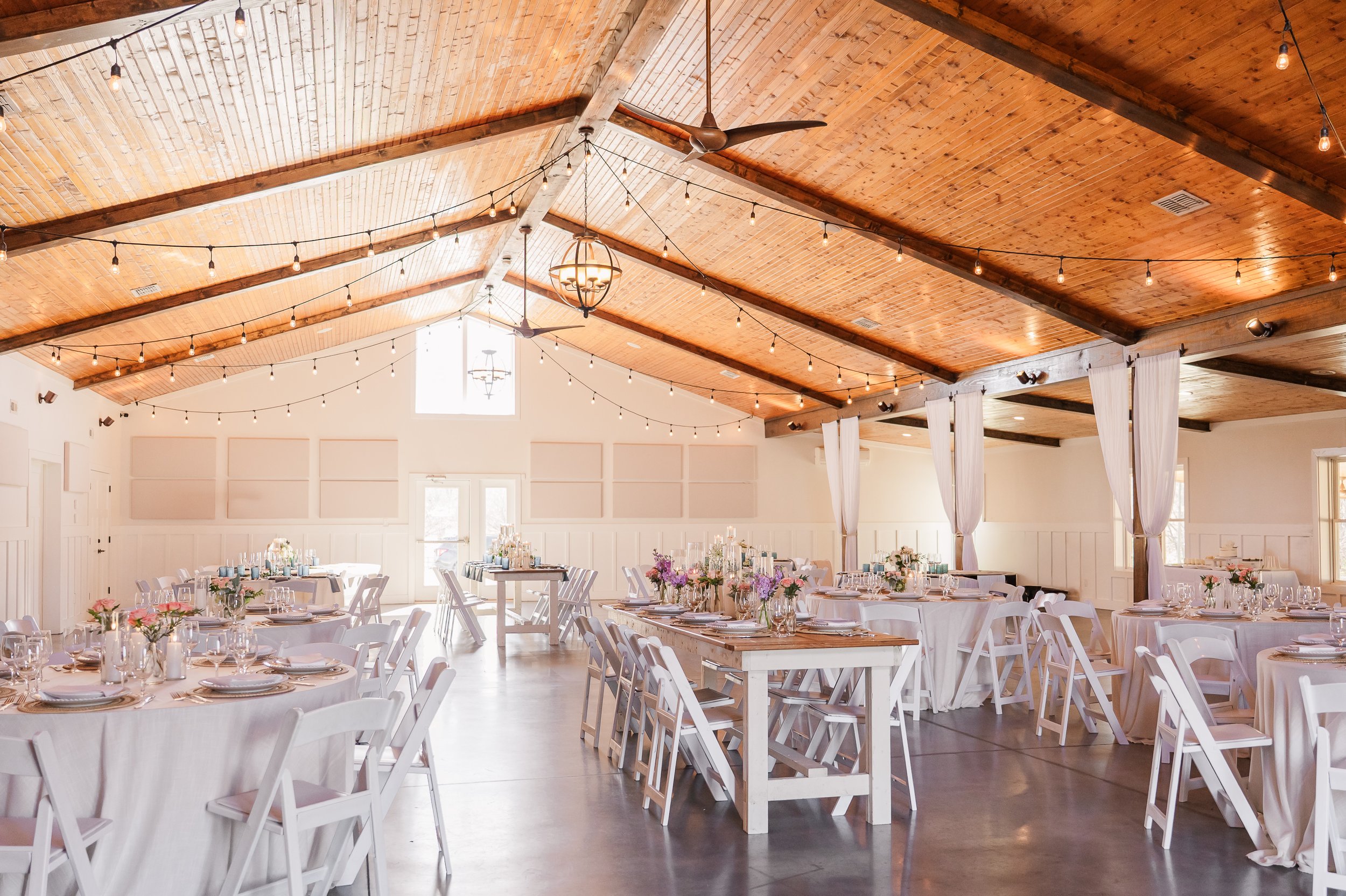Indoor event space with wooden vaulted ceiling, string lights, white tables with pastel tablecloths, and floral centerpieces.