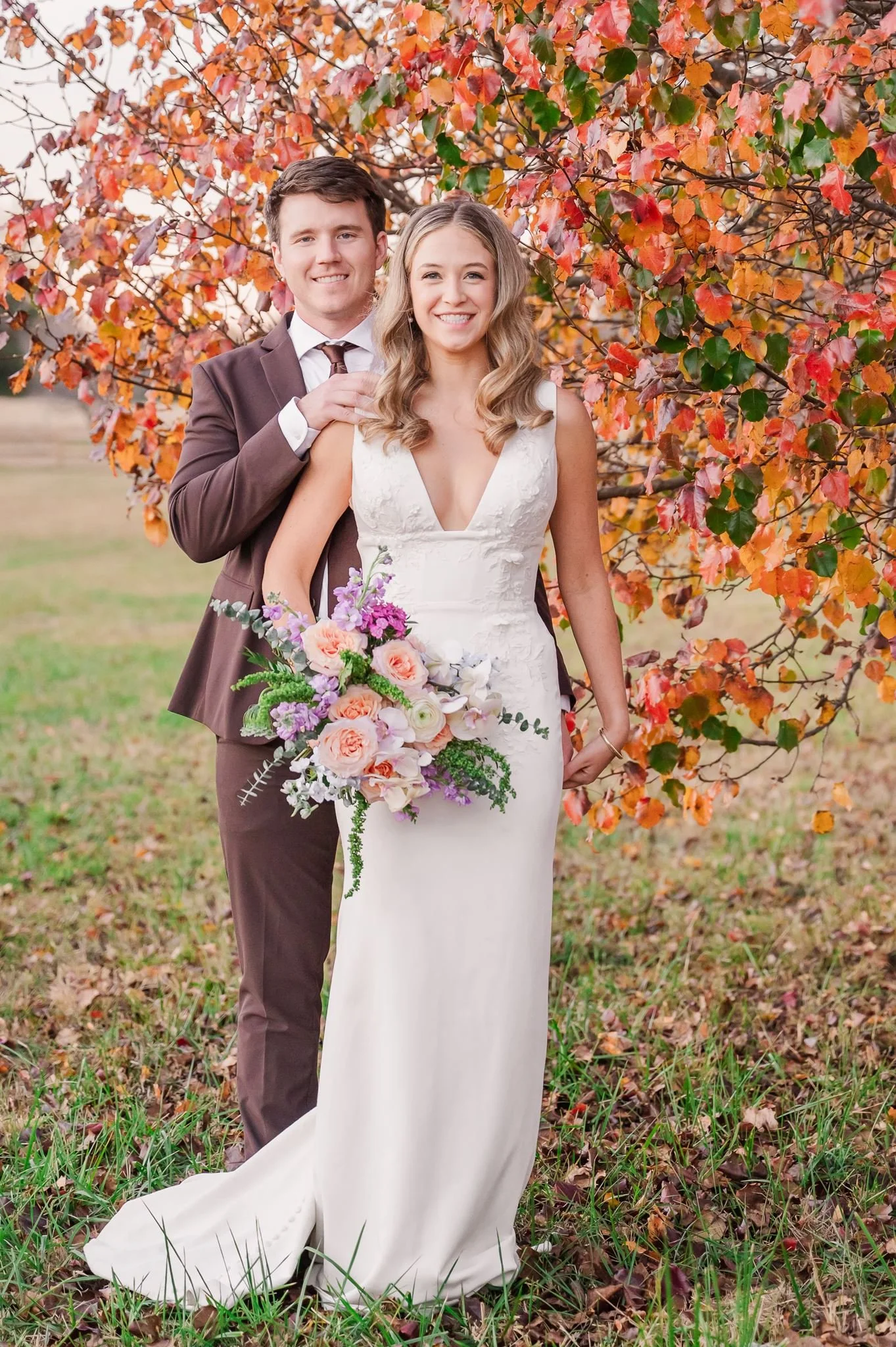 A happy couple in wedding attire standing outdoors in front of colorful autumn leaves, holding a bouquet of pink, purple, and white flowers, with the woman in a white dress and the man in a brown suit.