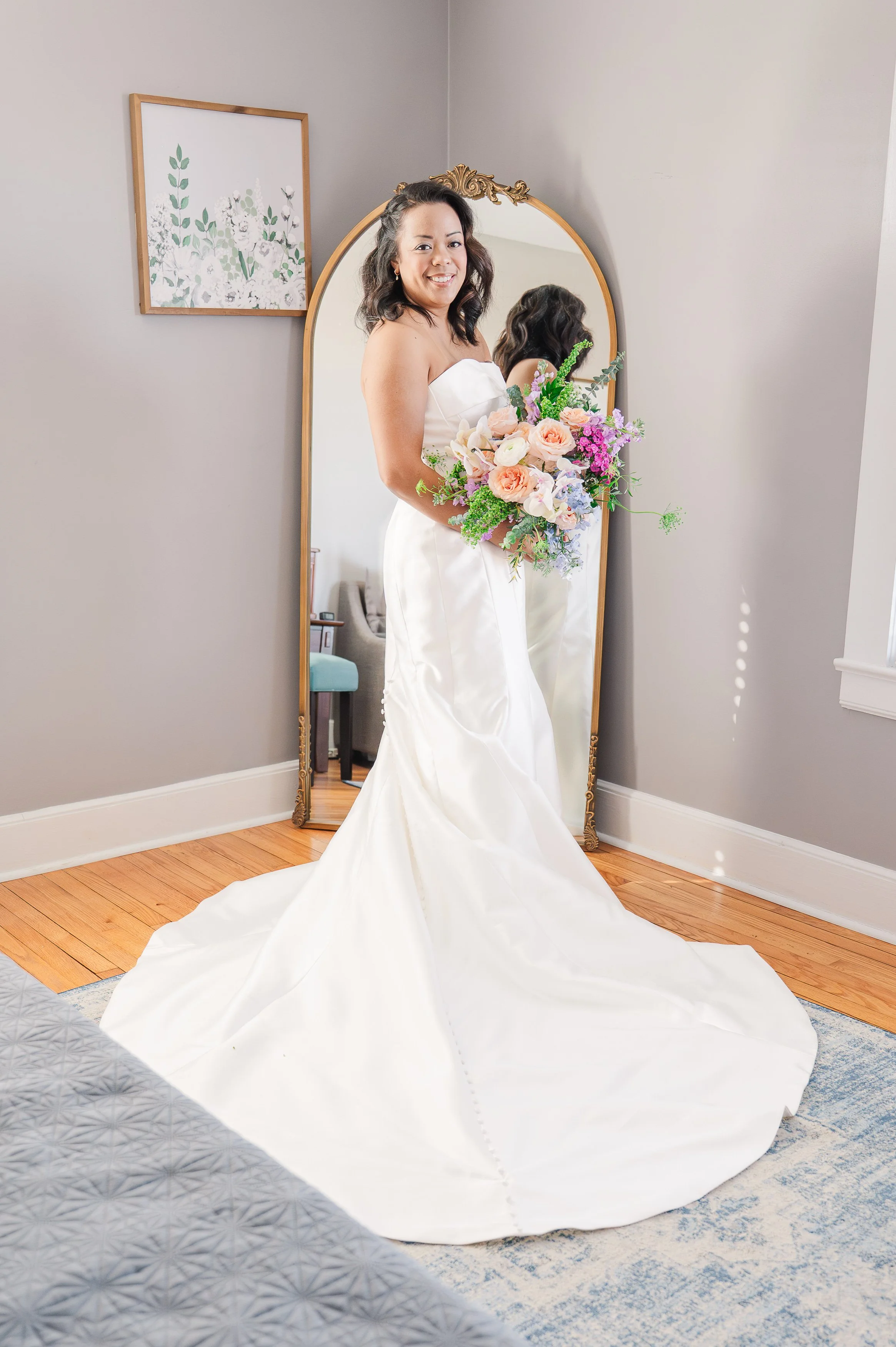 A woman in a white wedding dress holding a colorful bouquet of flowers, standing in front of a mirror in a room.