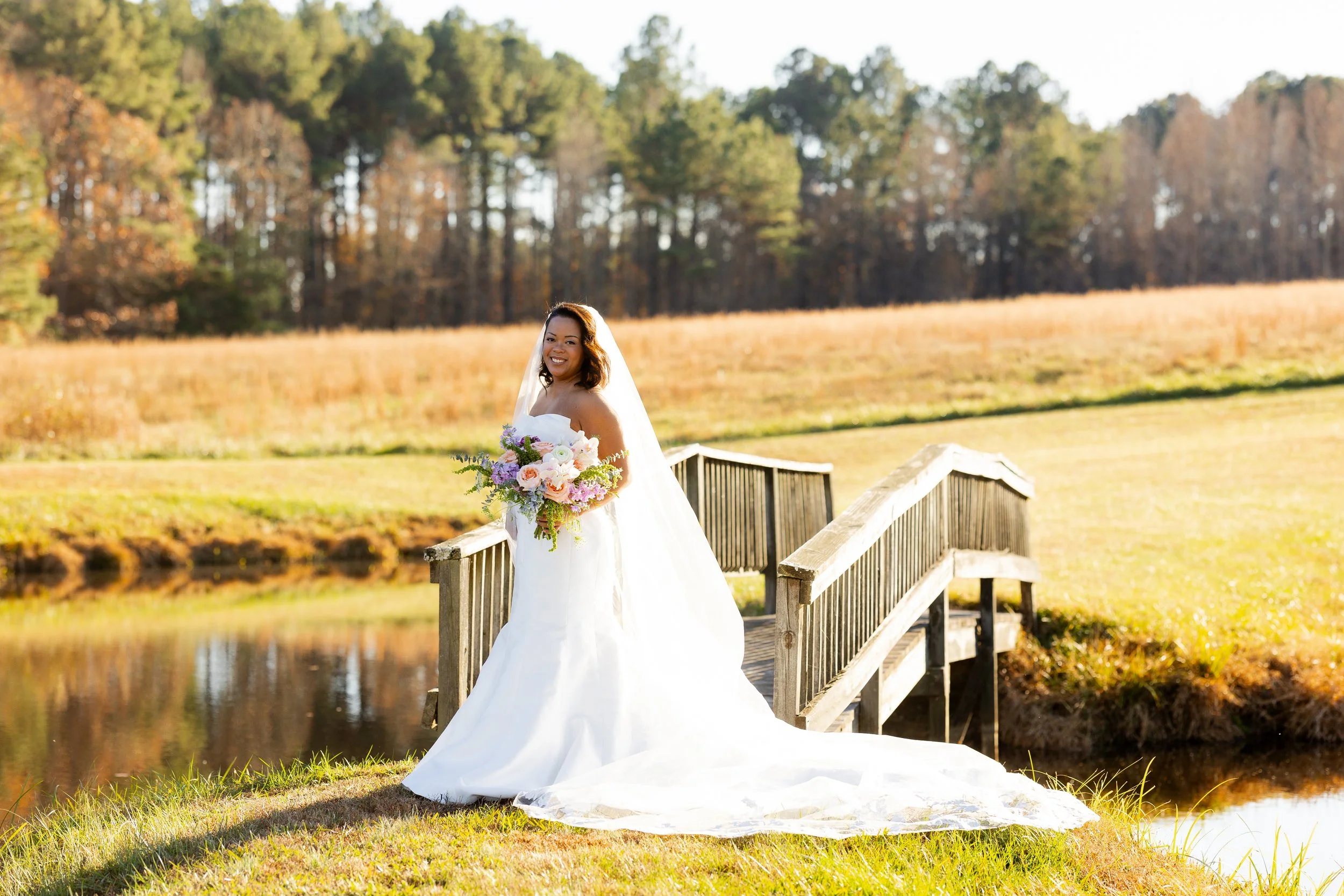 A bride in a white wedding dress holding a bouquet of flowers, standing on a small wooden bridge and smiling outdoors in a field with trees in the background.