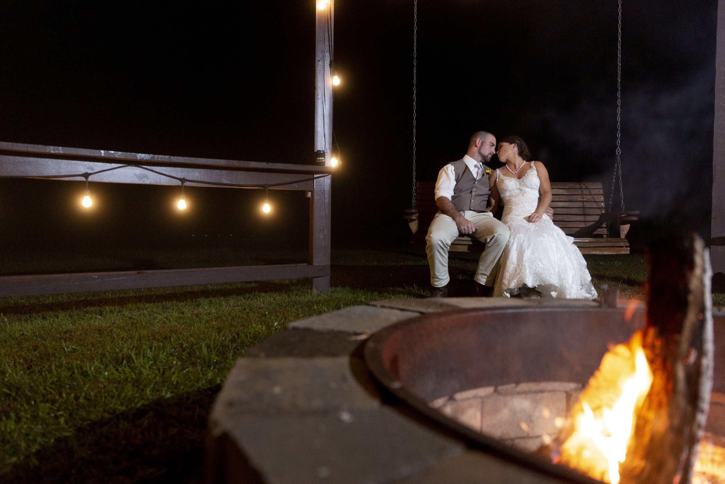 A bride and groom sitting close together on a wooden bench at night, illuminated by string lights, with a fire pit in the foreground.