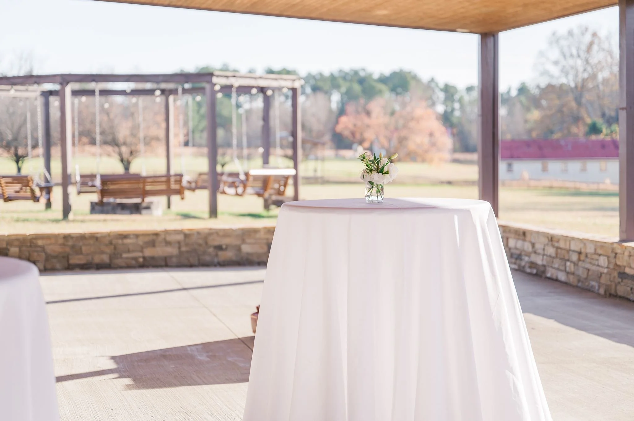 Outdoor patio area with a white round table, decorated with a glass vase of white flowers, and wooden benches and swings in the background under a wooden canopy, with trees and a house in the distance.