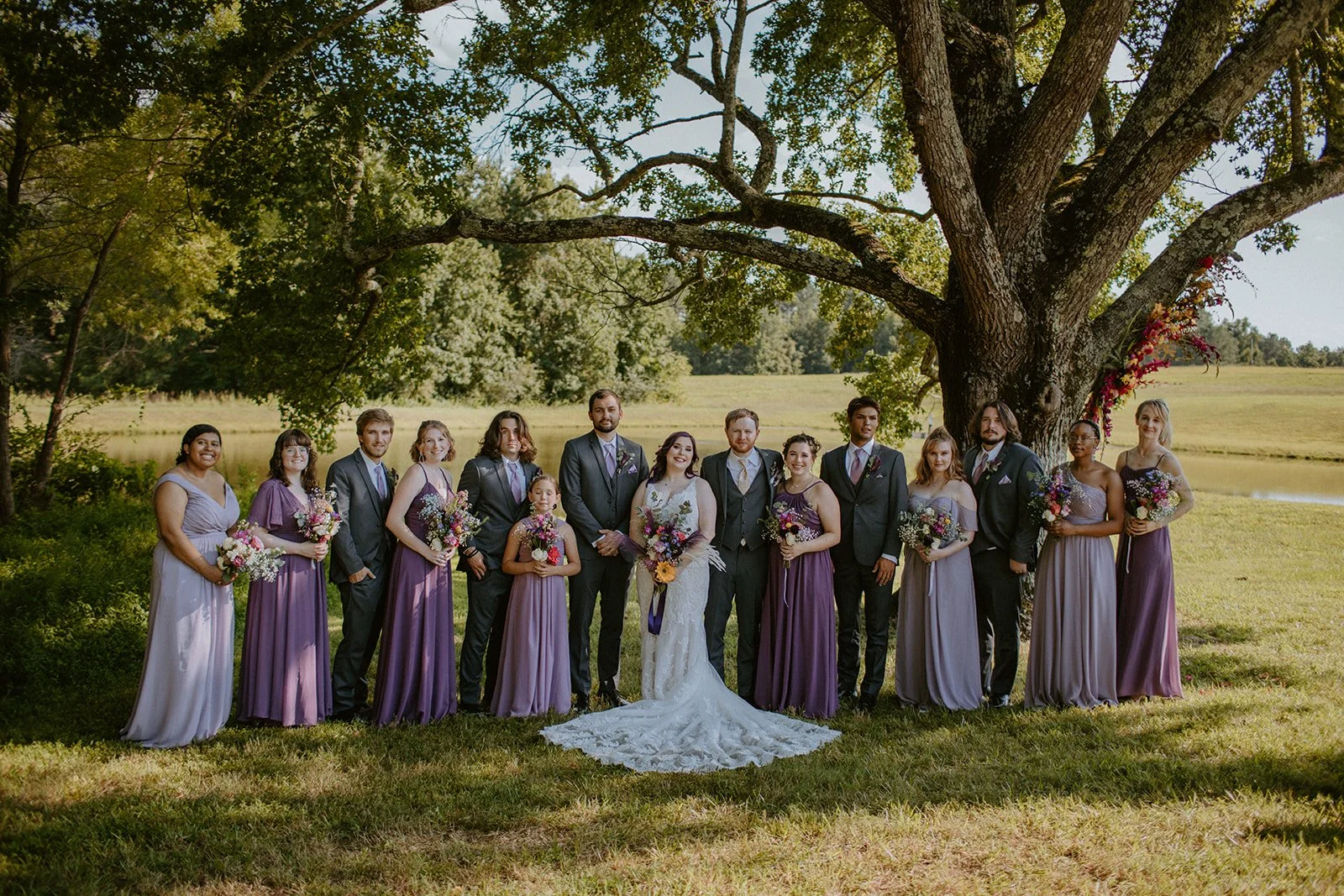A wedding party standing outdoors under a large tree, dressed in formal attire with the bride and groom at the center, all holding bouquets of flowers, with a field and trees in the background.