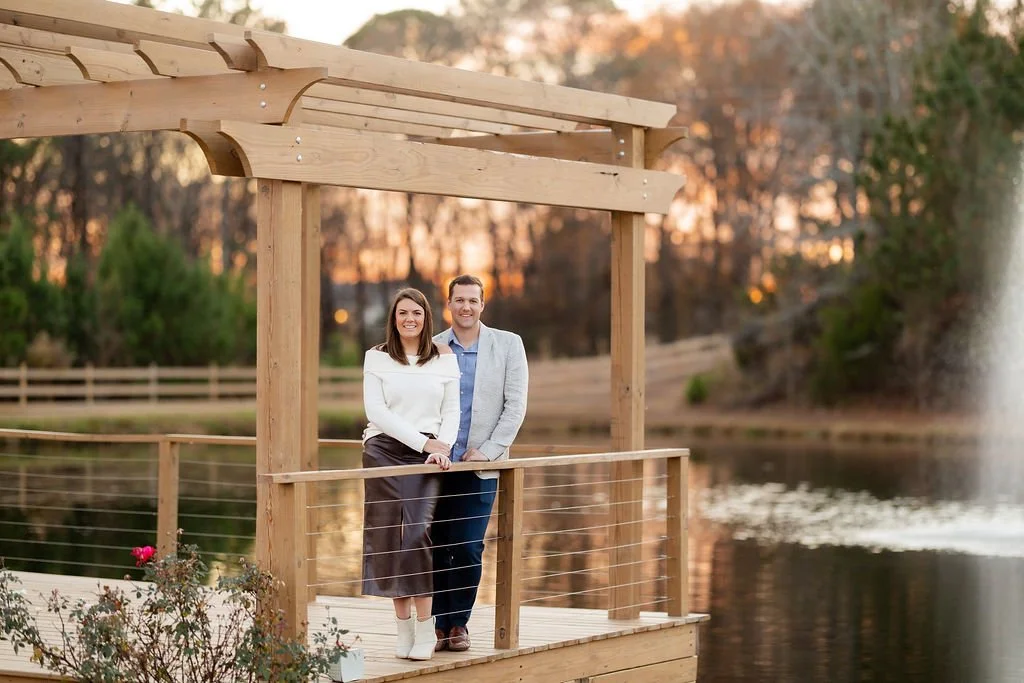 A smiling man in a light gray blazer and a smiling woman in a white dress with brown hair sitting on a wooden swing near a campfire during sunset.